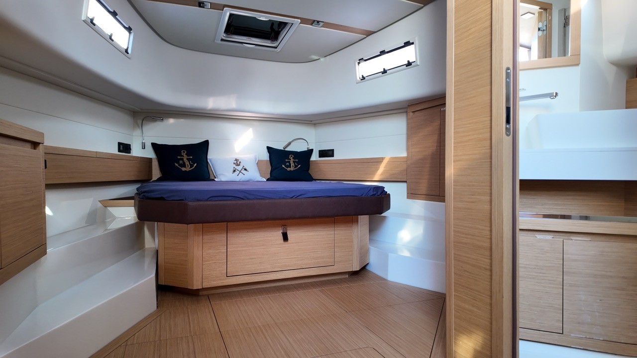 Pardo 43 motor yacht master cabin showing a large double berth with navy blue linen and navy anchor-embroidered cushions, light oak veneer throughout, under-berth storage drawers, overhead deck hatch and hull portlights for natural light, integrated reading lights, and open en-suite bathroom to the right