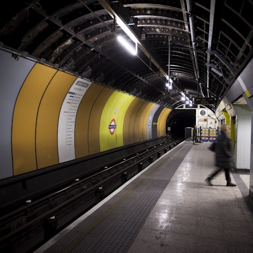 An empty underground train platform with yellow and white walls, overhead lights, and one person walking on the platform.