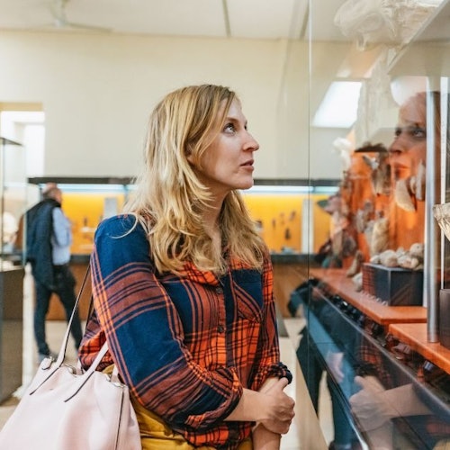 Mulher de camisa xadrez observando uma peça em uma vitrine de museu, com outros visitantes ao fundo.