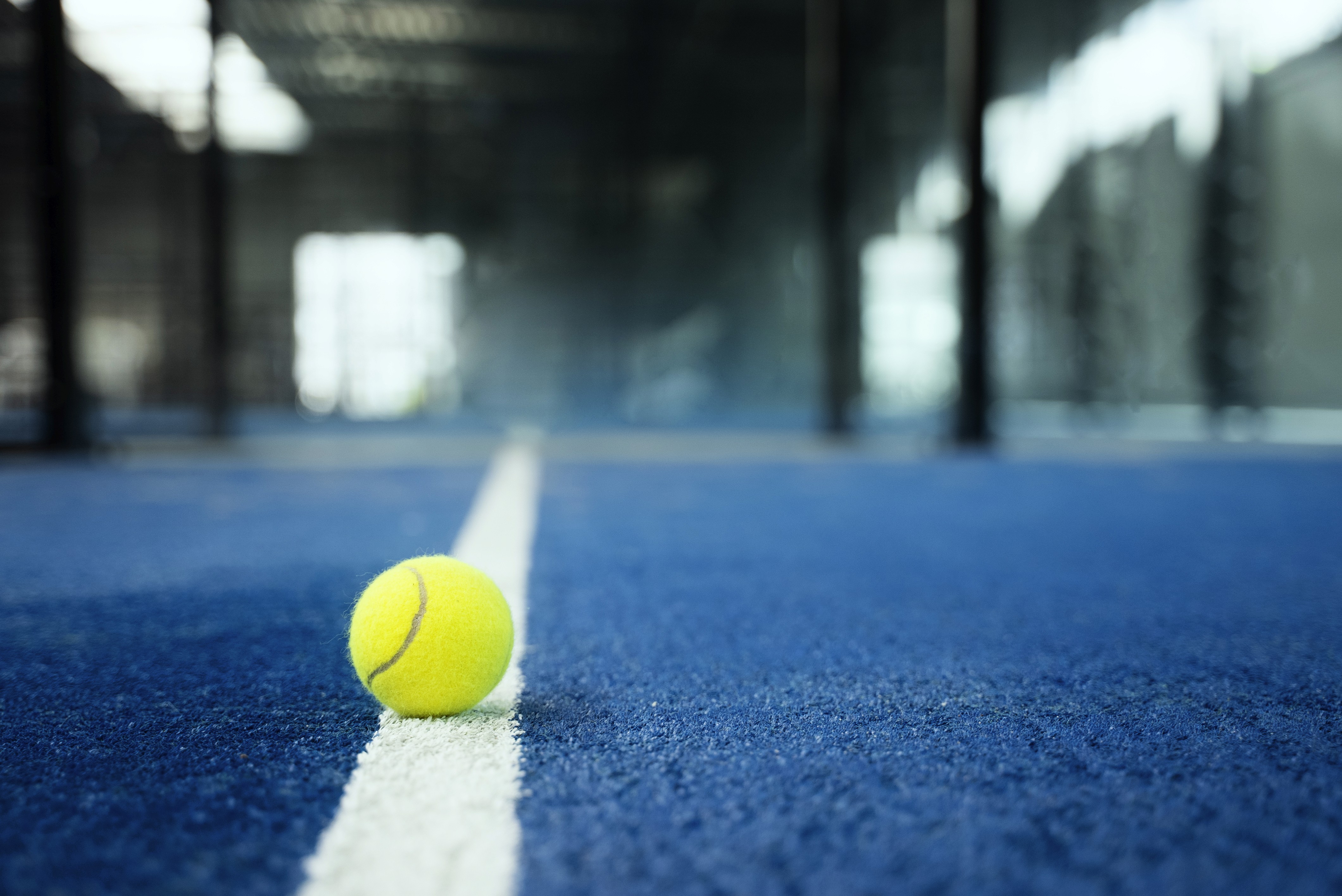 A yellow tennis ball rests on a vibrant blue court with a clear white line dividing the surface, set against the backdrop of an indoor sports facility with glass walls.