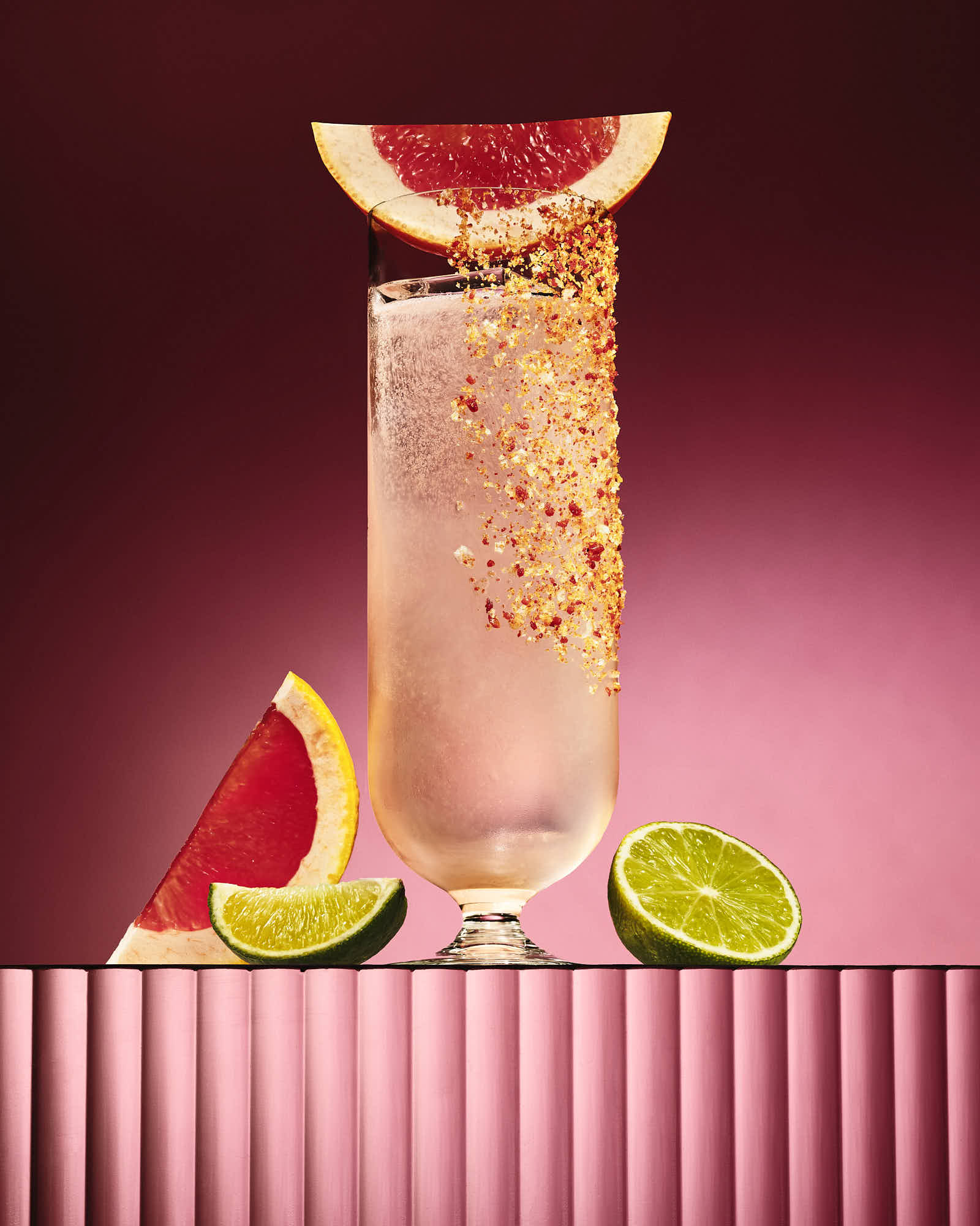 A tall glass of pink cocktail garnished with grapefruit slices, set against a vibrant pink background.