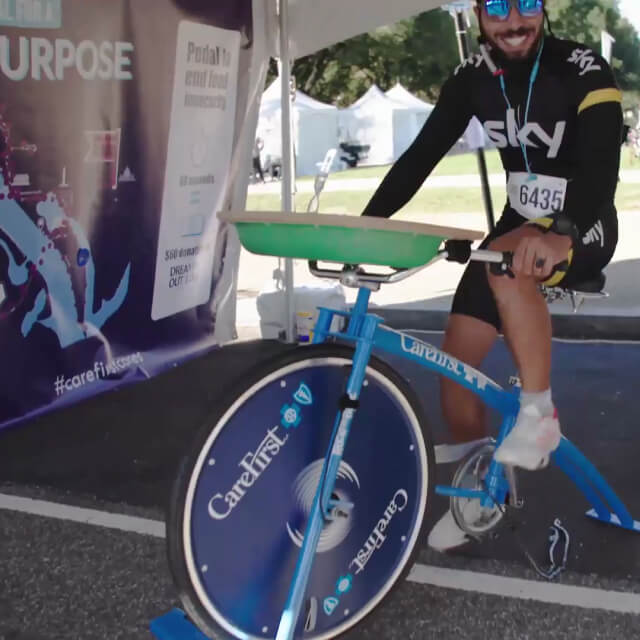 A person wearing athletic gear and sunglasses rides a stationary exercise bike at an outdoor event, with a tent and banners visible in the background.