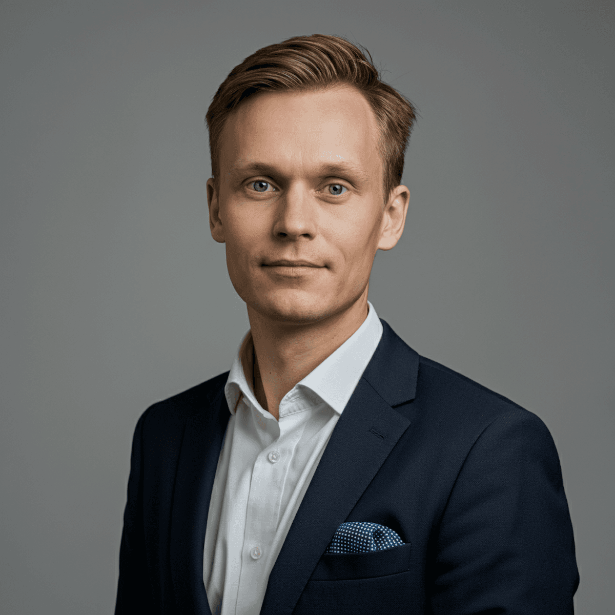 Professional headshot of a man in a dark suit and white shirt with a pocket square, facing the camera against a plain background.