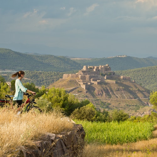 A person with a bicycle stands in a grassy field, looking at a distant hilltop fortress surrounded by green mountains.