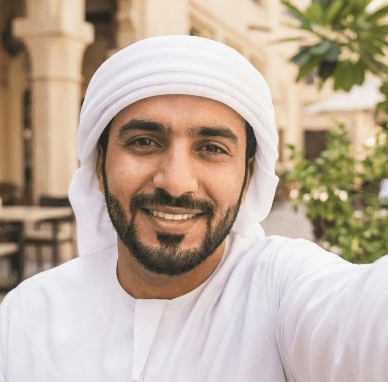 صورة سيلفي من زاوية عالية لرجل مبتسم ذو لحية داكنة ومهذبة، يرتدي كندورة بيضاء تقليدية وغترة بيضاء ملفوفة بأسلوب الحمدانية. يقف في ساحة خارجية مضيئة بأشعة الشمس تتميز بالعمارة التقليدية للشرق الأوسط، مع مبانٍ حجرية بلون الرمال، وأبواب مقوسة، وأشجار خضراء ذات أوراق في الخلفية.