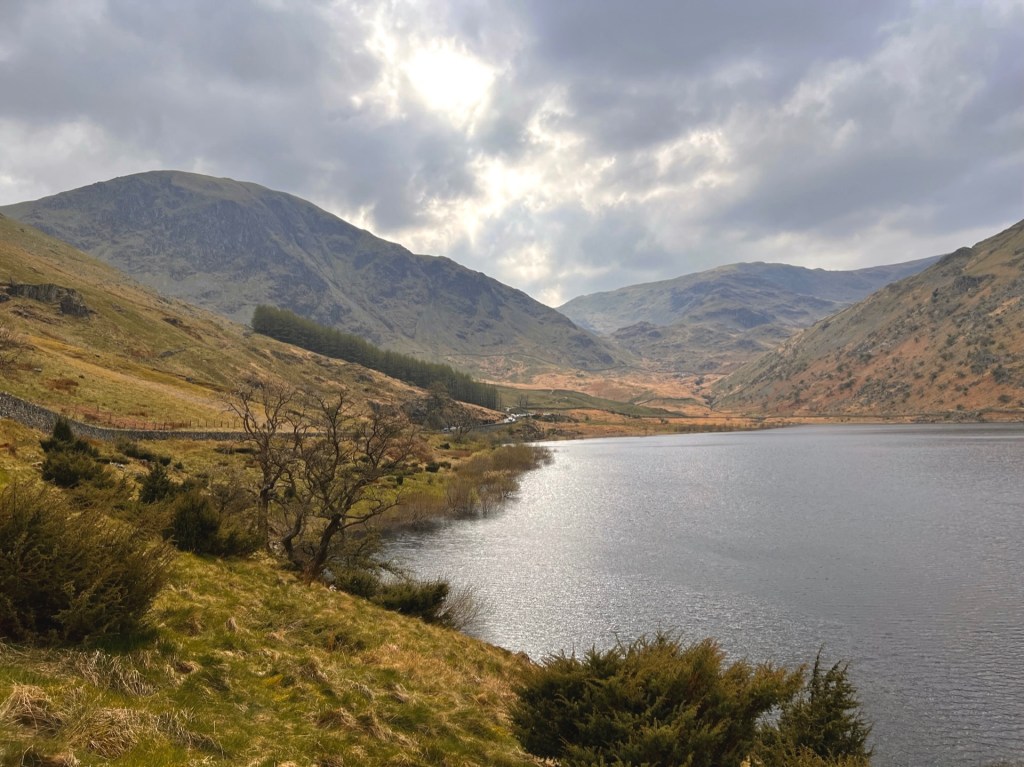 Haweswater reservoir on the right. A dry stone wall runs alongside the road on the left, leading to a car park. Mountains in the background.