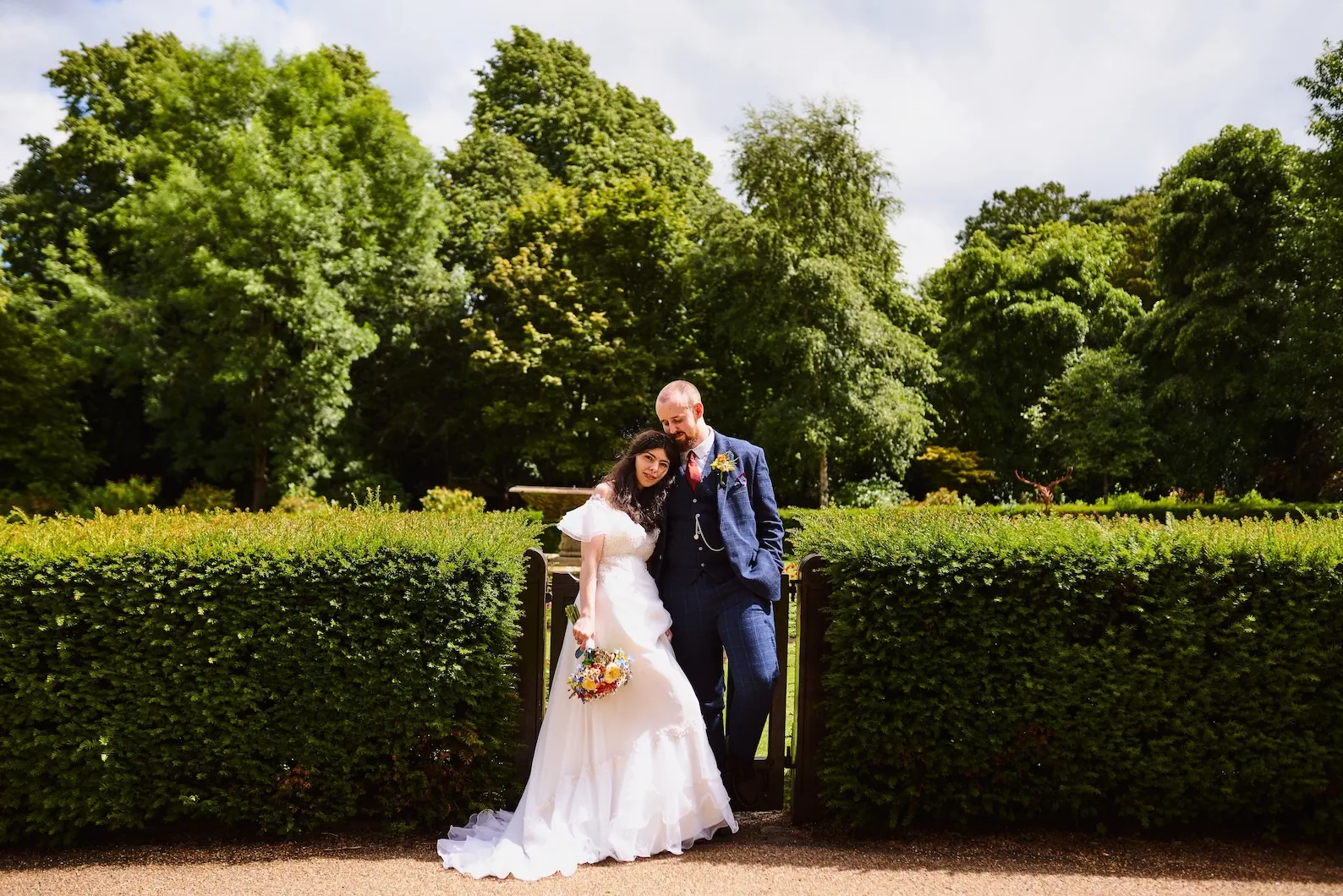A bride in a white gown leaning against her groom in a navy checked suit, in front of a hedge and tall green trees