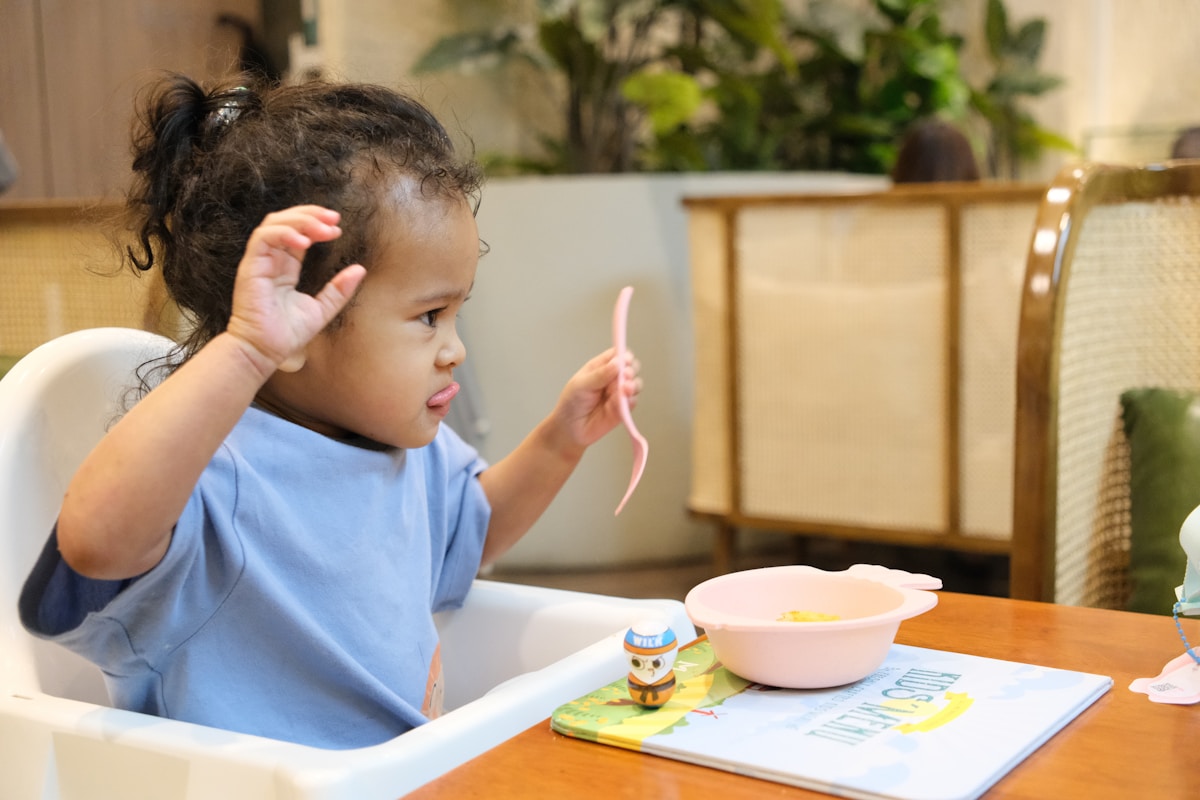 A young child sitting in a high chair at the table holding a spoon and looking at a bowl of food