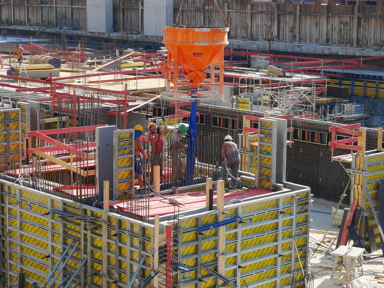 Construction site with workers in hard hats setting up formwork for a concrete pour. An orange concrete pump is in use. Busy, industrial atmosphere.