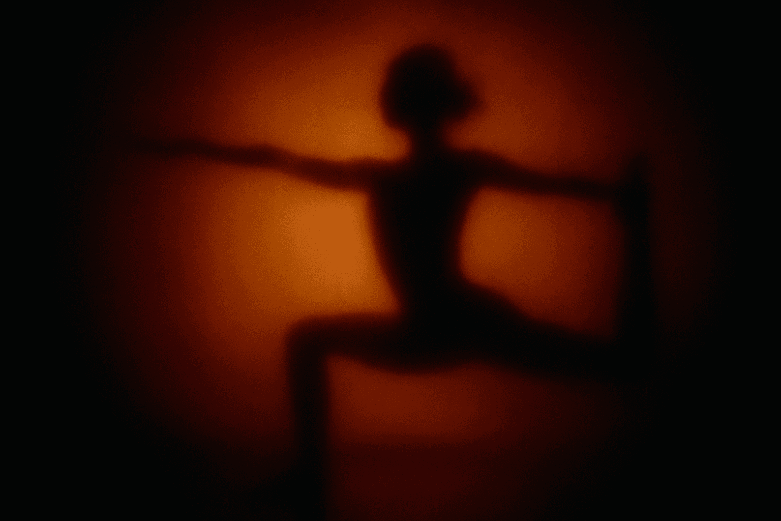 The image shows a dark silhouette of a person in a yoga stretching pose set against a vibrant orange background. The person appears to be in a balancing position with one arm extended forward and the other hand reaching back to hold the foot, likely in a dancer's pose or similar stretch. The soft focus and strong backlighting create a dramatic and artistic shadow effect, highlighting the contours and posture of the figure. This image can be used to represent topics related to yoga, fitness, mindfulness, meditation, and physical health.