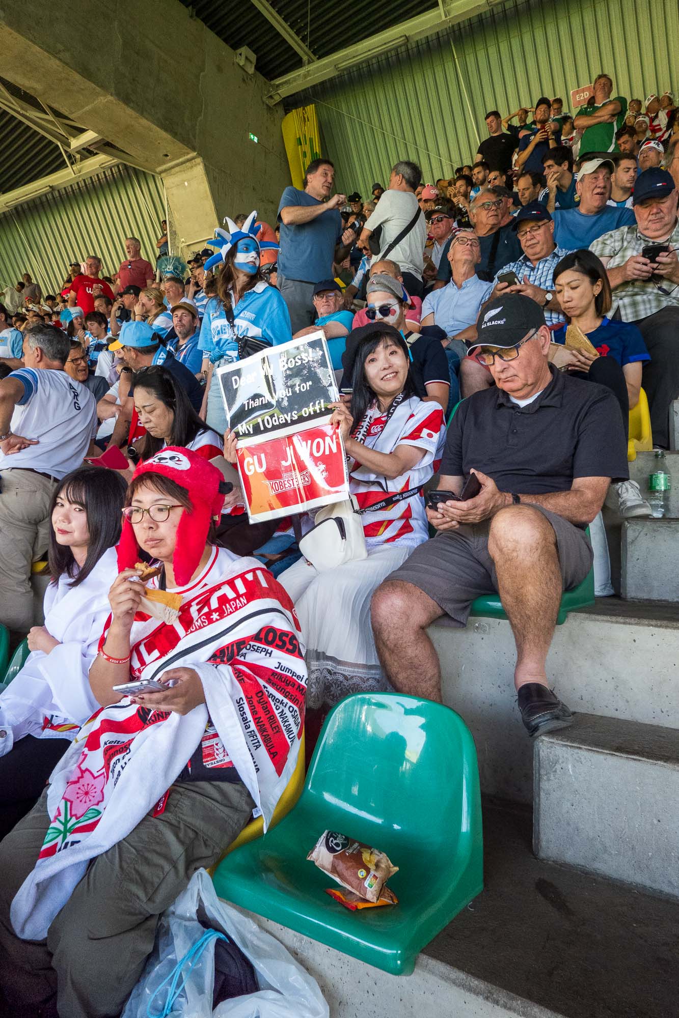 Supportrice japonaise brandissant une pancarte « Dear my boss! Thank you for my 10 days off! », entourée de supporters japonais, d’un homme à la casquette All Blacks et d’une Argentine au visage peint