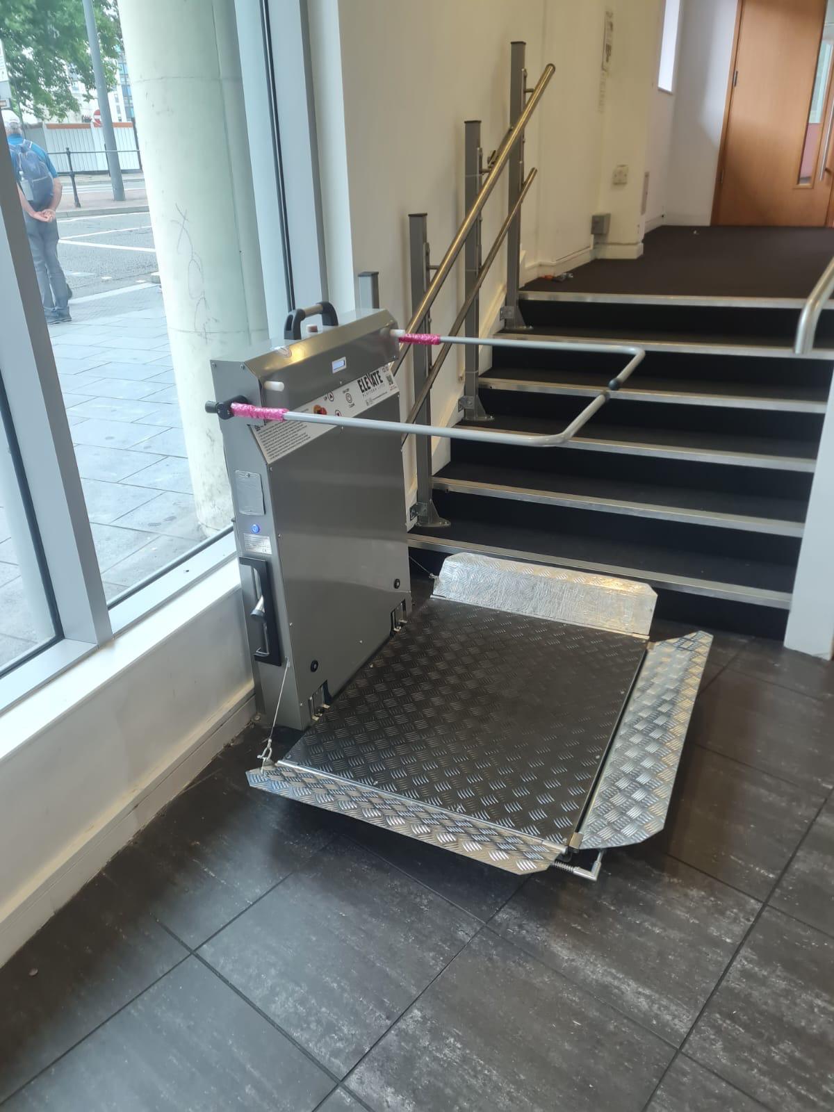 Straight incline platform stairlift at lower landing with safety arms raised and platform partially folded — stainless steel drive unit beside commercial staircase with glazed frontage
