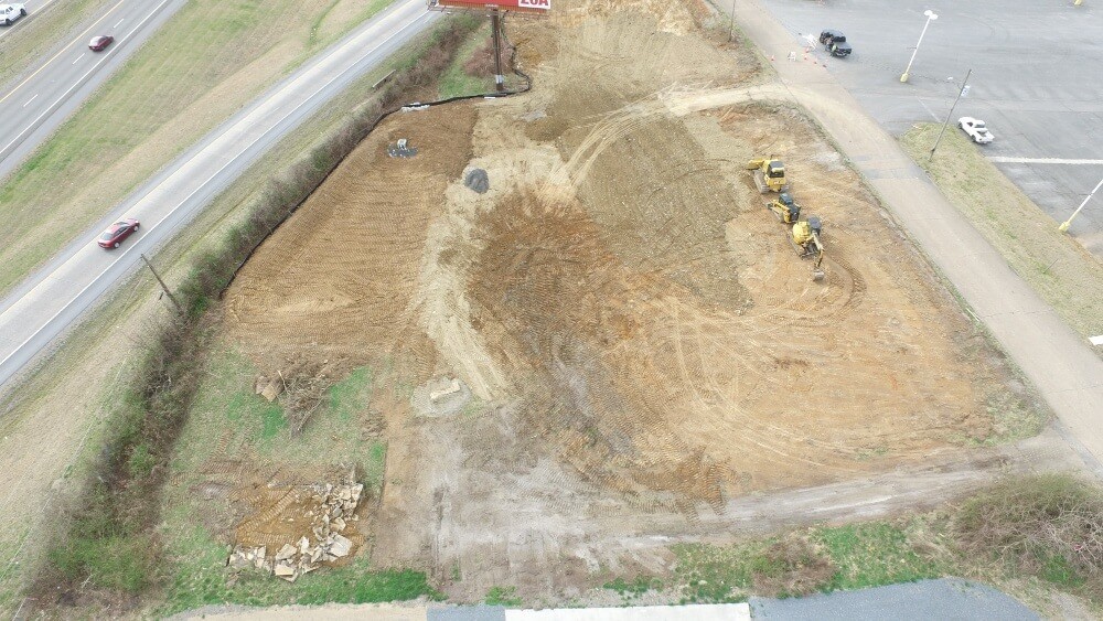 High altitude drone shot of a vacant lot being graded with highway on one side and parking lot on the other