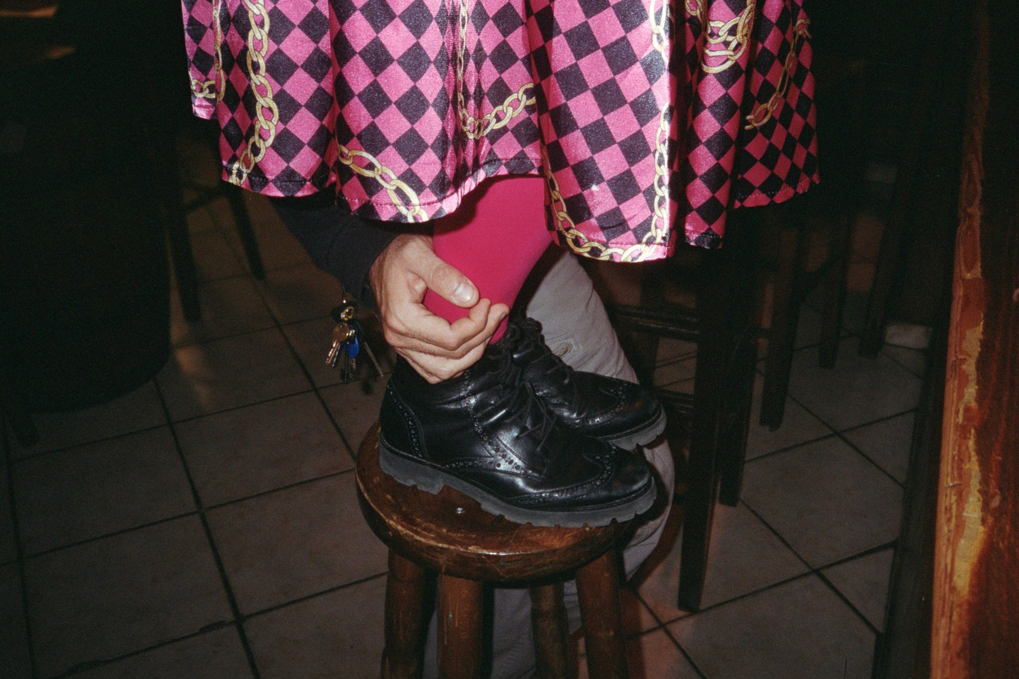 A person wearing a pink and black checkered skirt with gold chains, bright pink tights, and shiny black shoes is sitting on a wooden stool in a tiled room.