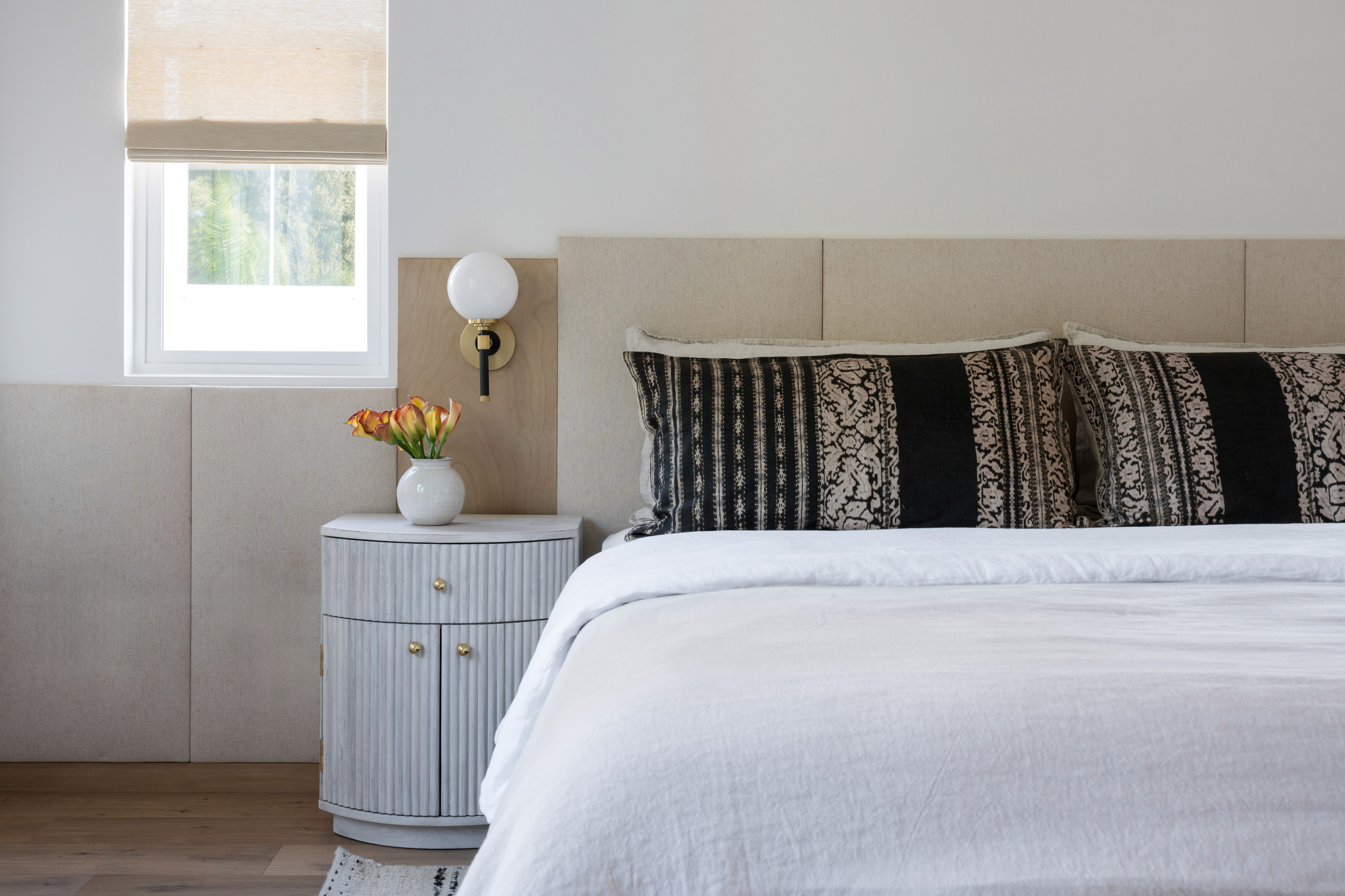 Bright bedroom with white linen bedding, patterned lumbar pillows, globe wall sconce, and curved nightstand by Jubilee Interiors in Arcadia, California