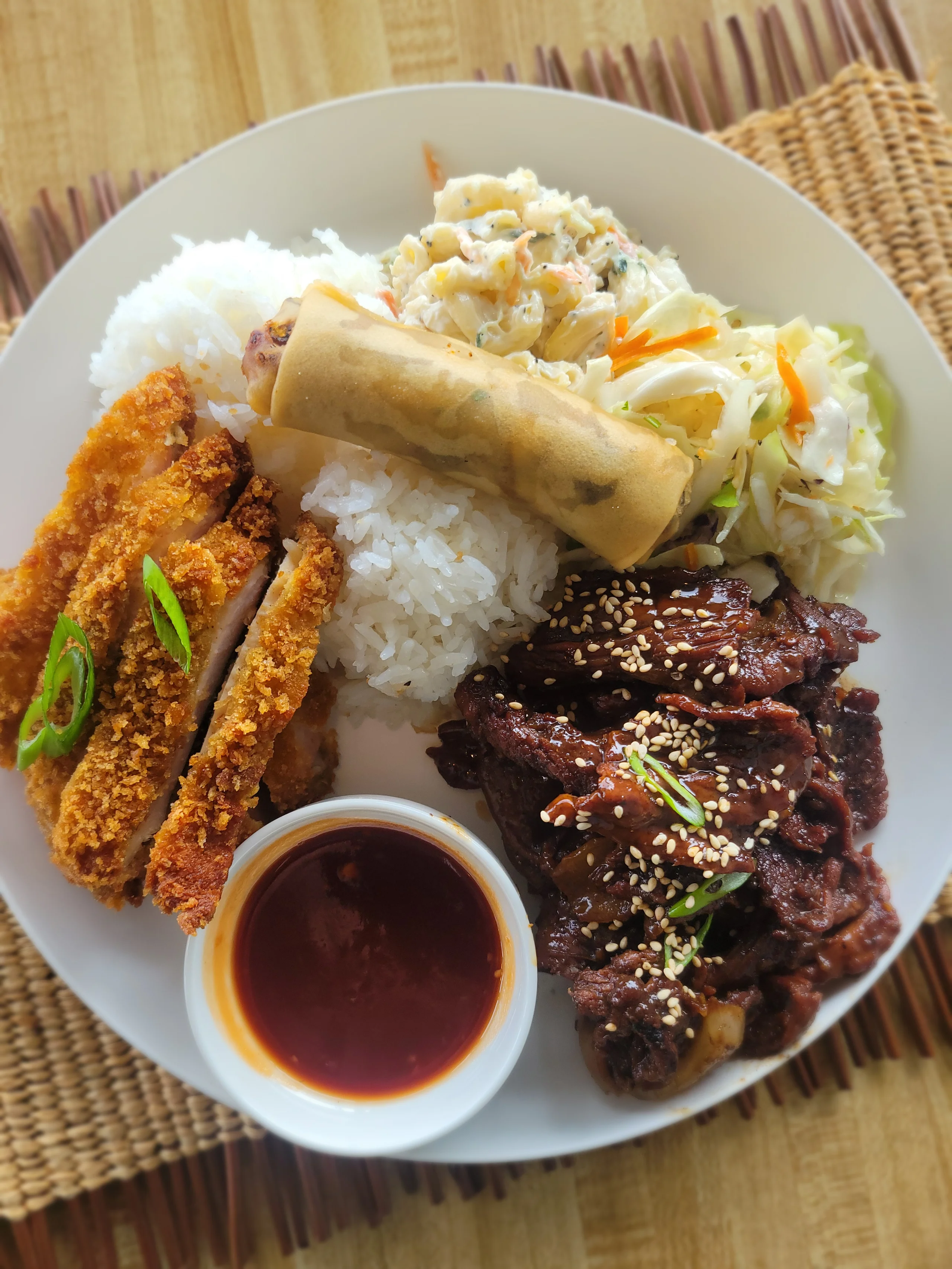 A large white plate featuring thin-sliced teriyaki beef and golden-fried chicken katsu strips, served with a spring roll, white rice, macaroni salad, and Asian slaw.