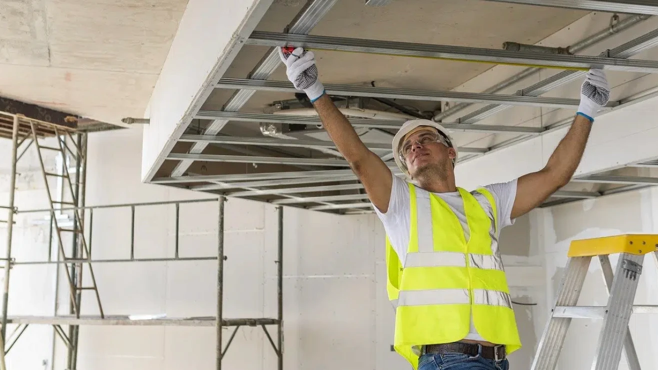 Person wearing a white helmet, safety goggles, gloves, and a yellow reflective vest, working on the installation of a suspended metal structure in an environment under construction.