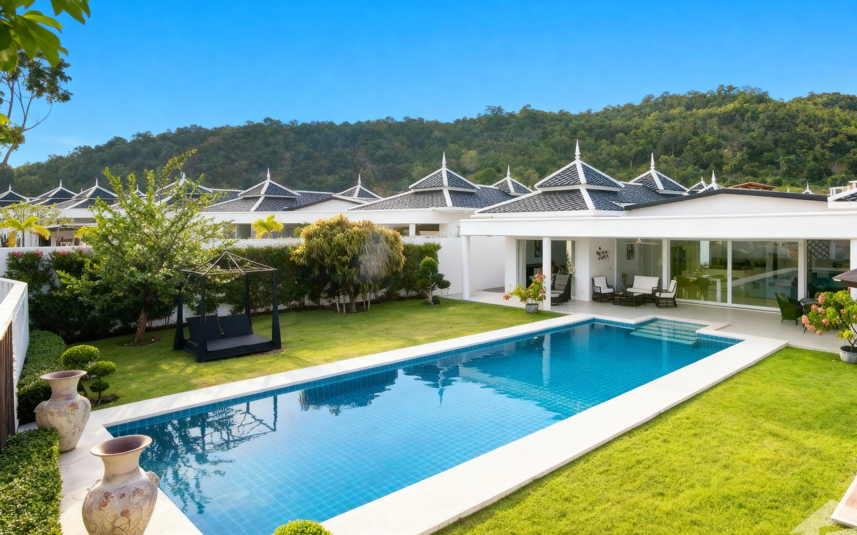 Modern family-friendly villas in Soi 102/112, South Hua Hin, with beige facades, glass balconies, and private pools, surrounded by green lawns and a palm tree under a cloudy sky.