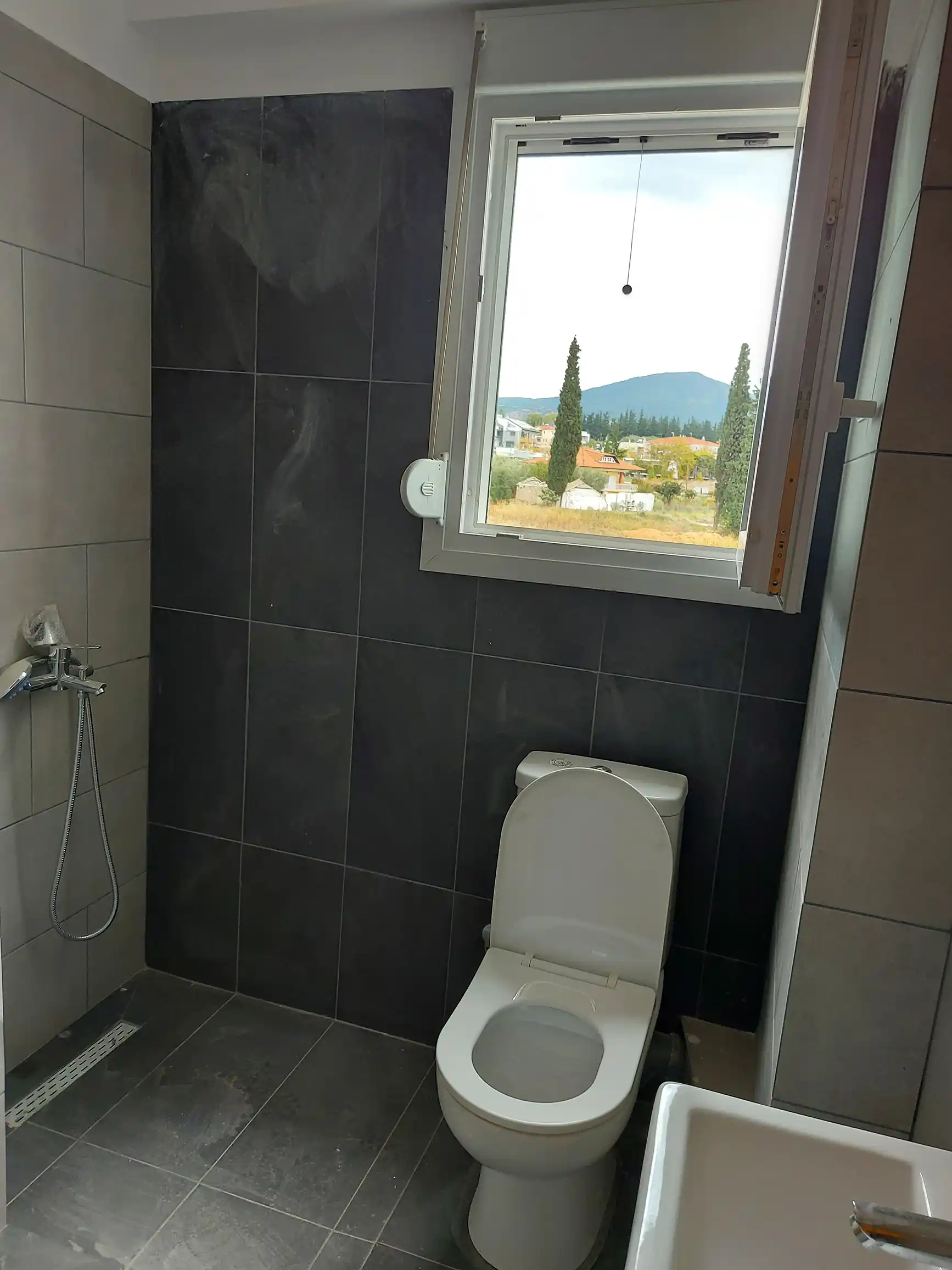 Μοντέρνο μπάνιο με σκούρα γκρι πλακάκια, λευκή λεκάνη και παράθυρο με όμορφη θέα στο βουνό.