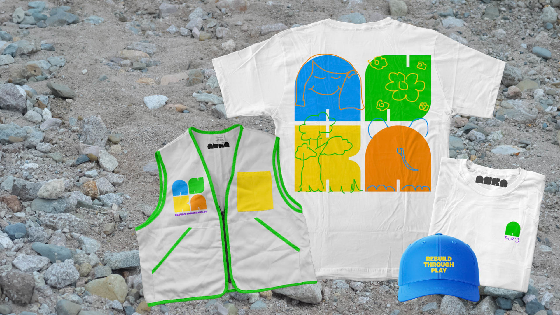 A white sleeveless vest with green trim and a yellow square patch is positioned on a rocky surface. Beside it are two white t-shirts: one with four colorful abstract designs in blue, green, yellow, and orange, and the other with a green logo and a small green leaf logo. A blue baseball cap with the text "BUILD THROUGH PAY" and a green logo is visible. The background consists of gray rocks and pebbles.