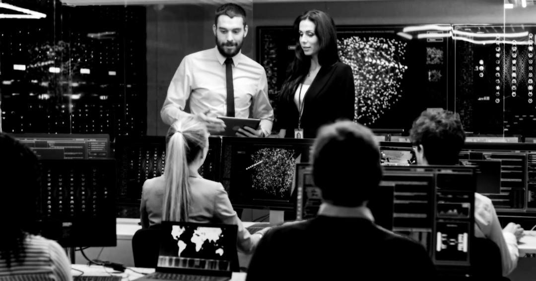 Black and white photo of an incident response room. Screens with graphs on the background. Team of 4 researchers are working on computers. 2 team leaders are  standing, facing them, man and woman, coordinating the team. 