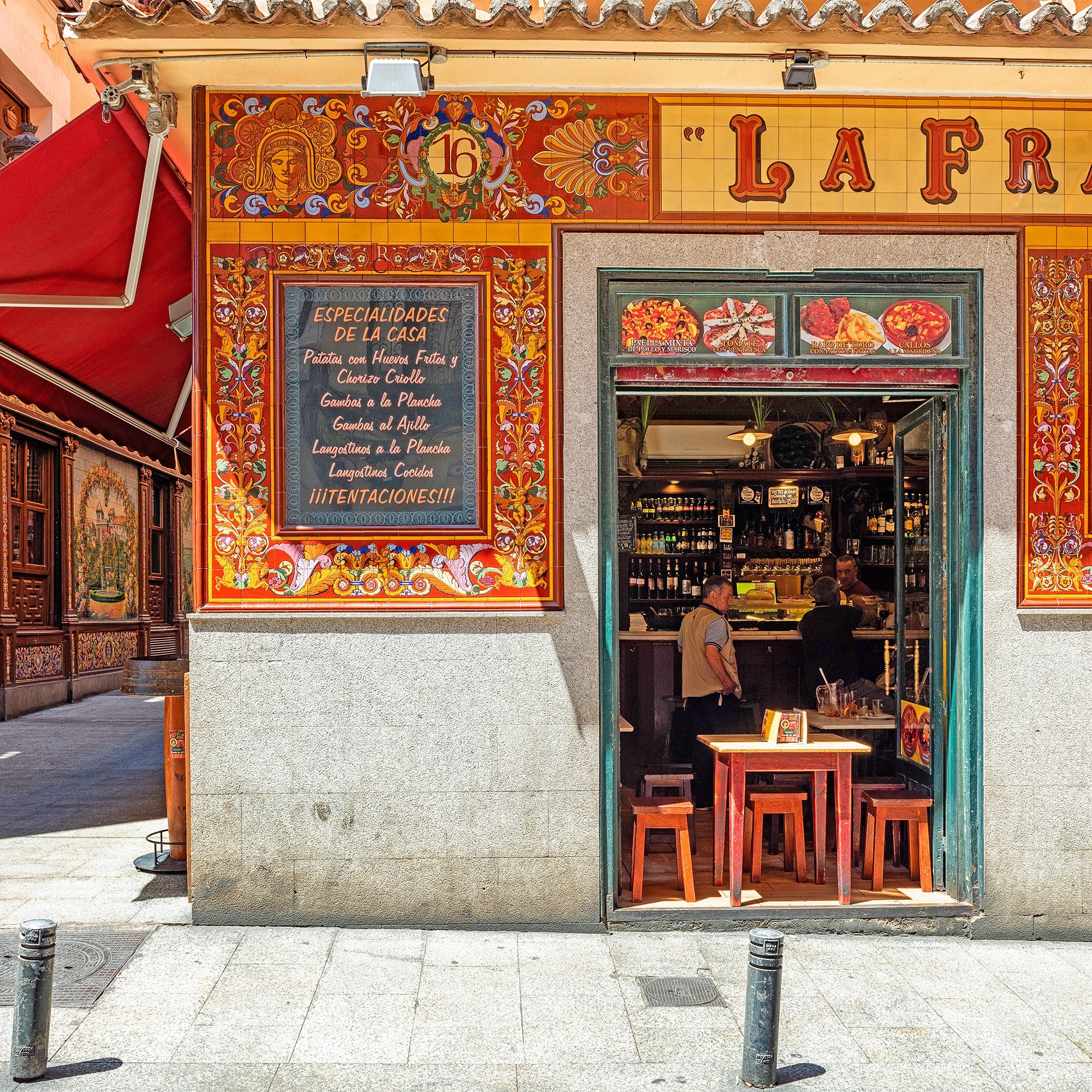 A tiled tapas taberna in Madrid, its azulejo façade above red stools set out on the pavement.