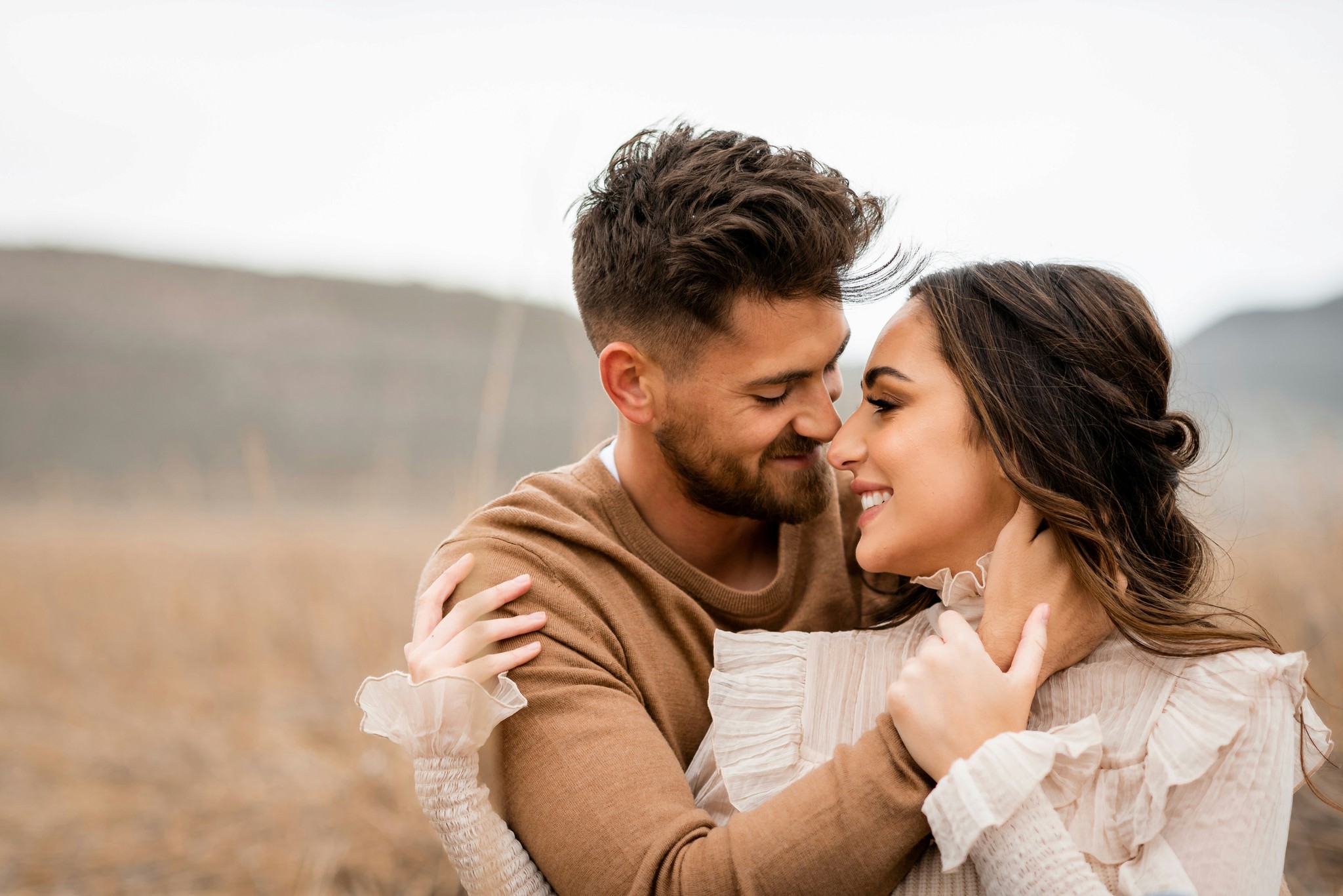 Close-up of a smiling couple touching foreheads in a warm embrace during an engagement session in a windswept open field