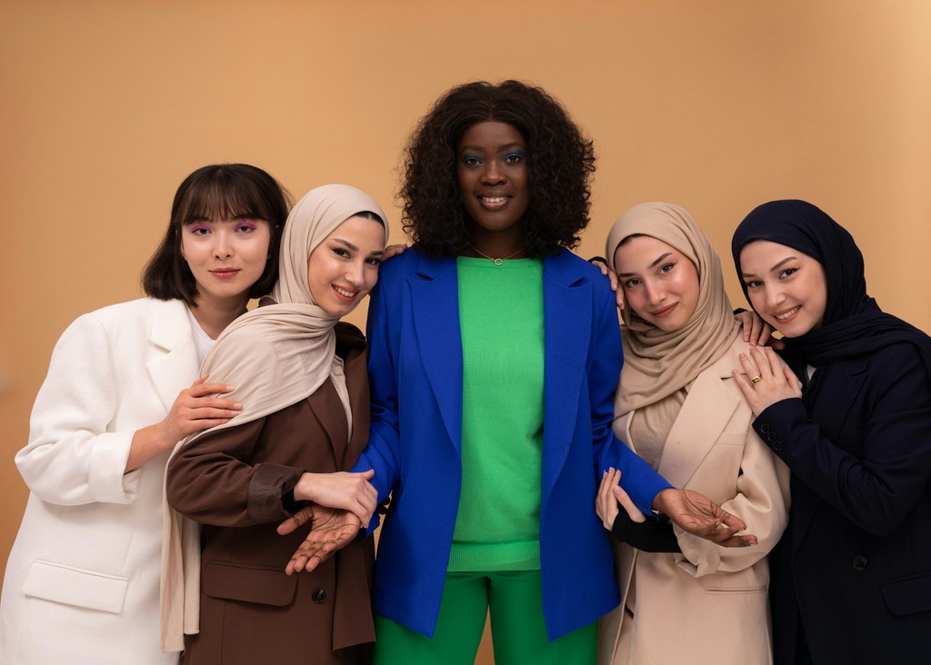 A diverse group of five women, dressed in stylish and modern attire, pose together against a beige background. The women, showcasing different cultures and styles, exude confidence and unity, with one woman in a vibrant blue blazer and green outfit standing in the center, while the others wear various hijabs and fashionable outfits, smiling warmly.