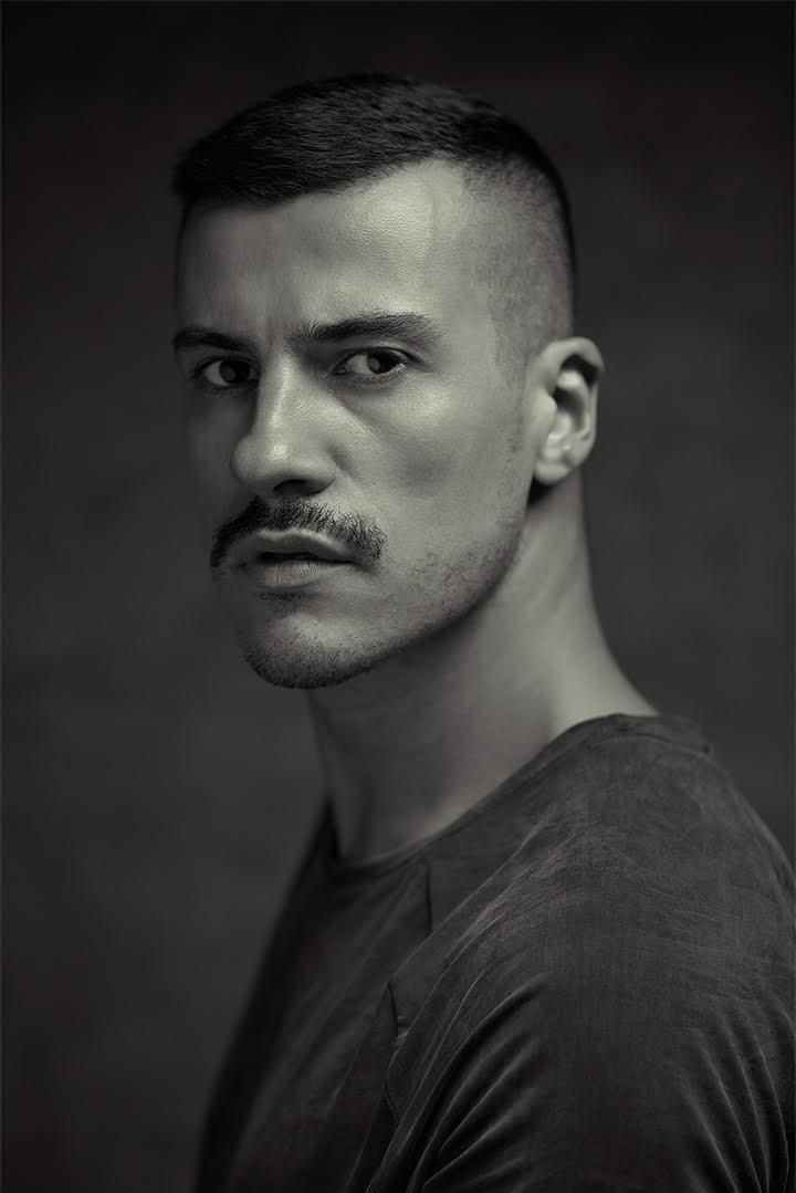 A confident person with short, styled hair and a leather jacket poses against a dark backdrop.