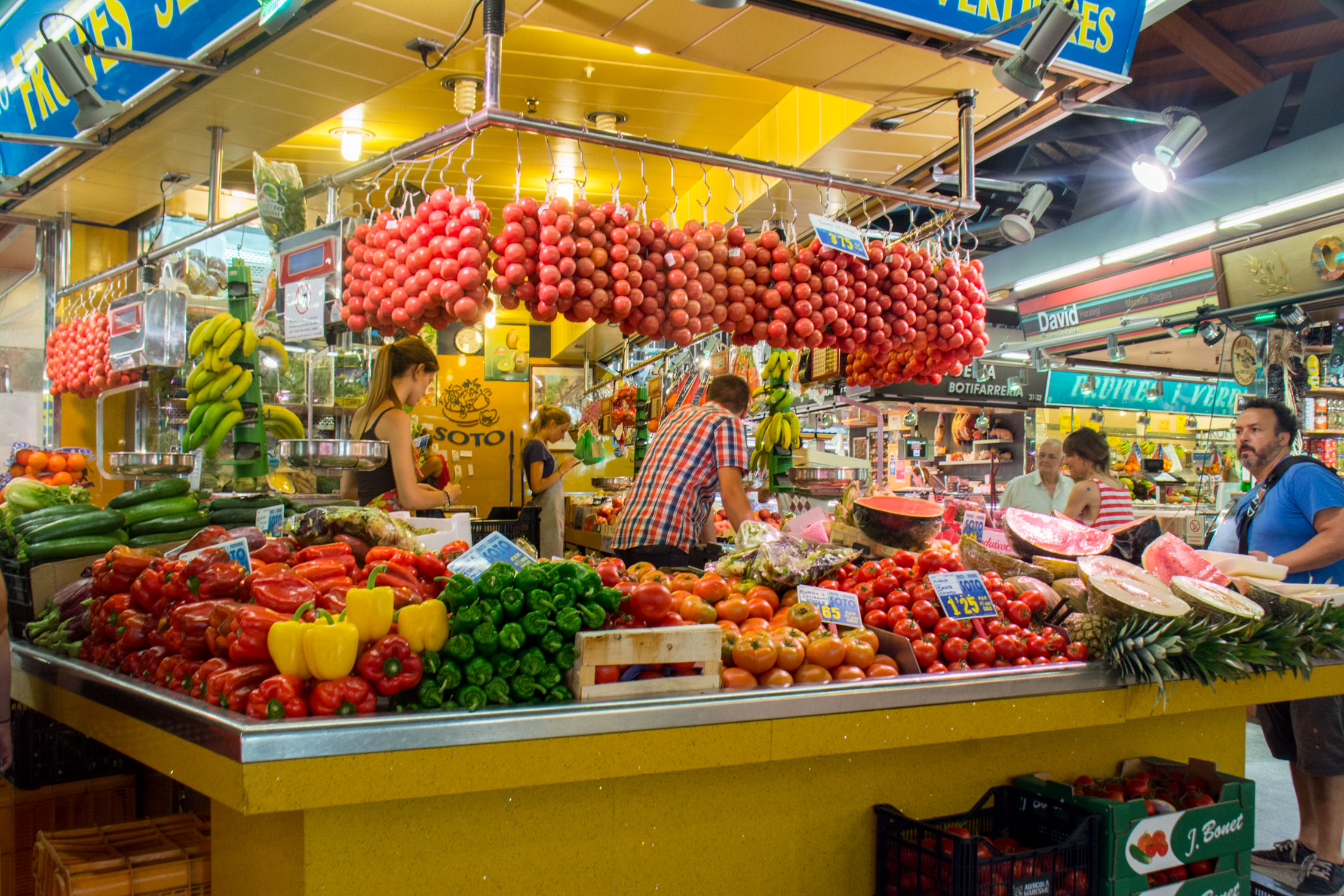 Vibrant stalls overflow with colorful fresh produce under bright indoor lighting in a bustling market setting.