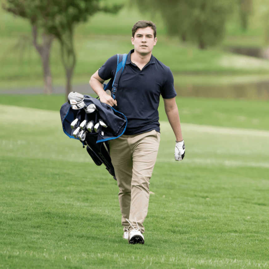 A golfer walks across a green field carrying a golf bag over his shoulder, dressed in casual golf attire.
