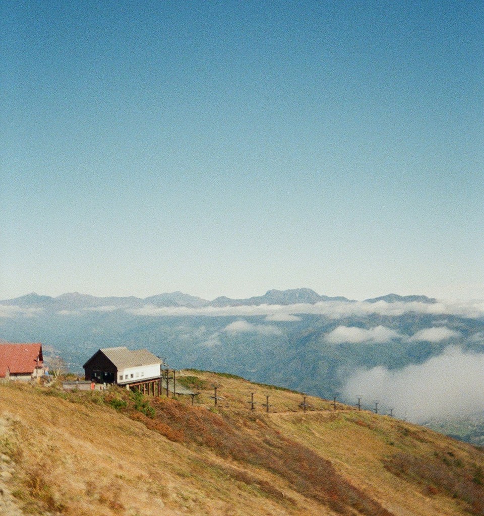 Photograph of a mountainside with a small wooden lodge perched on a grassy slope, overlooking a valley filled with low clouds and distant peaks. Soft, hazy light and muted colors create a tranquil, atmospheric landscape.