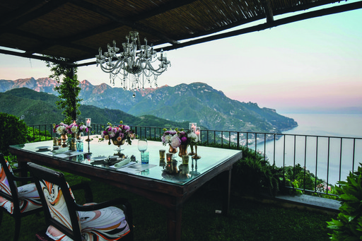A scenic view from a balcony overlooking the ocean at sunset, with a dining table set nearby.