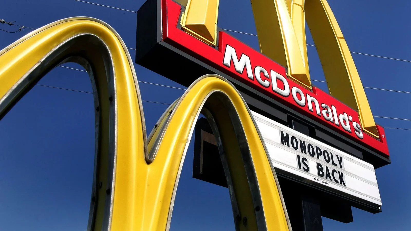 McDonald's golden arches sign with "McDonald's" text and "MONOPOLY IS BACK" marquee against blue sky.