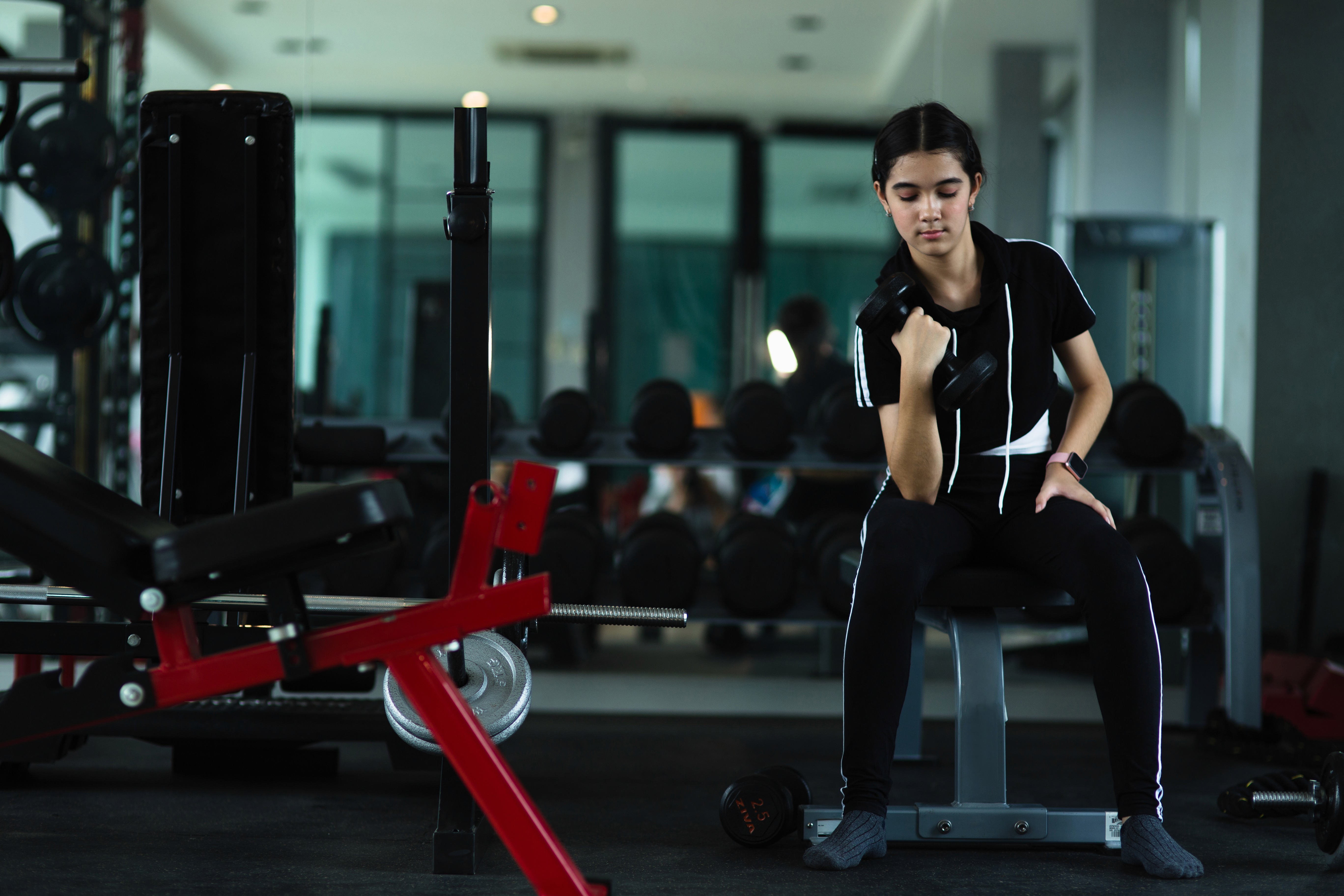 A young woman sitting on a gym bench performing a bicep curl with a dumbbell. She is dressed in black athletic wear, with her hair tied back, and appears focused on her exercise. The gym setting around her features various equipment, such as weight benches, dumbbell racks, and mirrors, creating a functional workout environment. The lighting is soft, highlighting her calm determination as she exercises. The overall mood conveys focus and dedication during a strength training session.