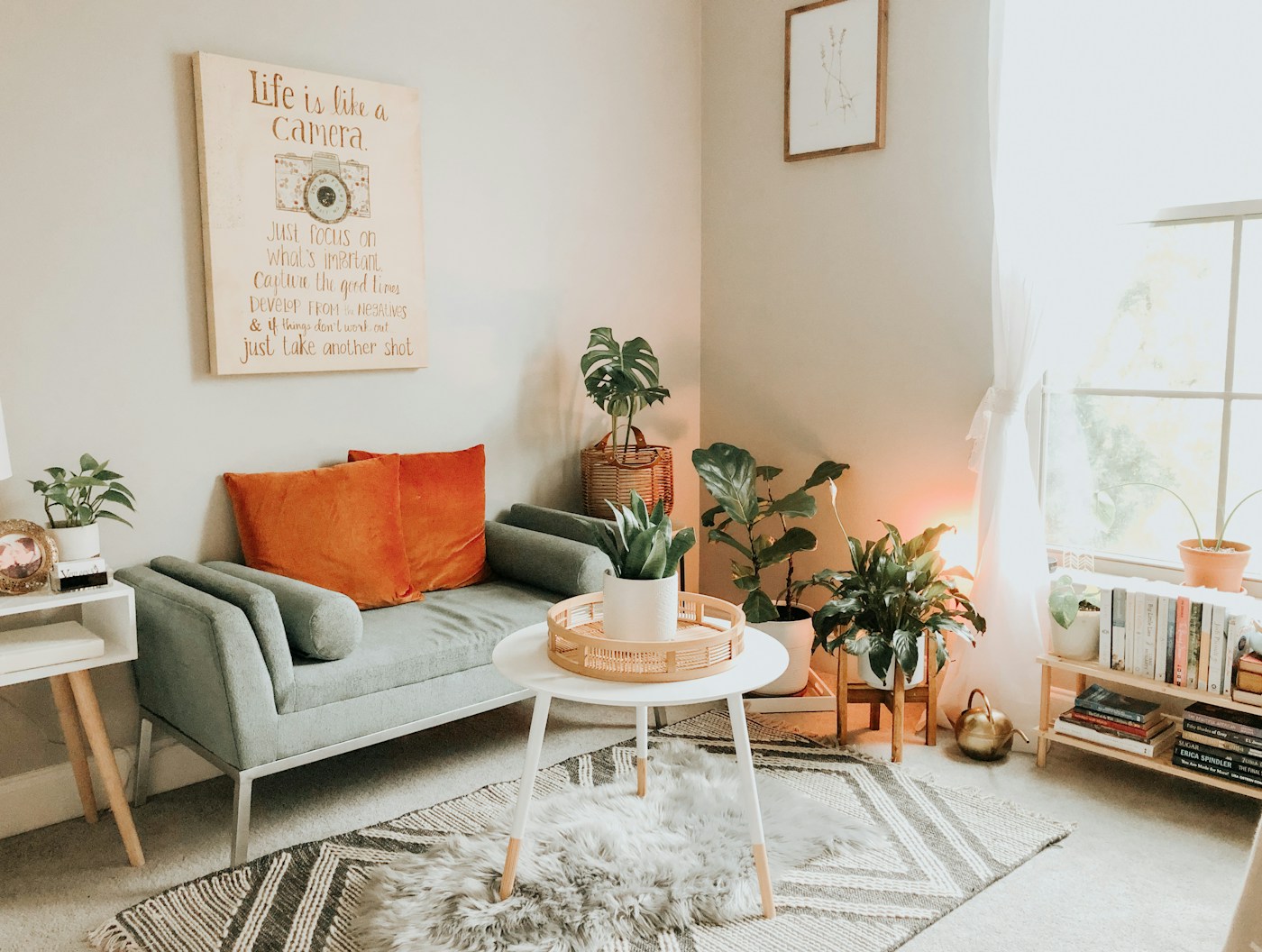 A sage sofa with orange cushions, a round coffee table and plants by the window