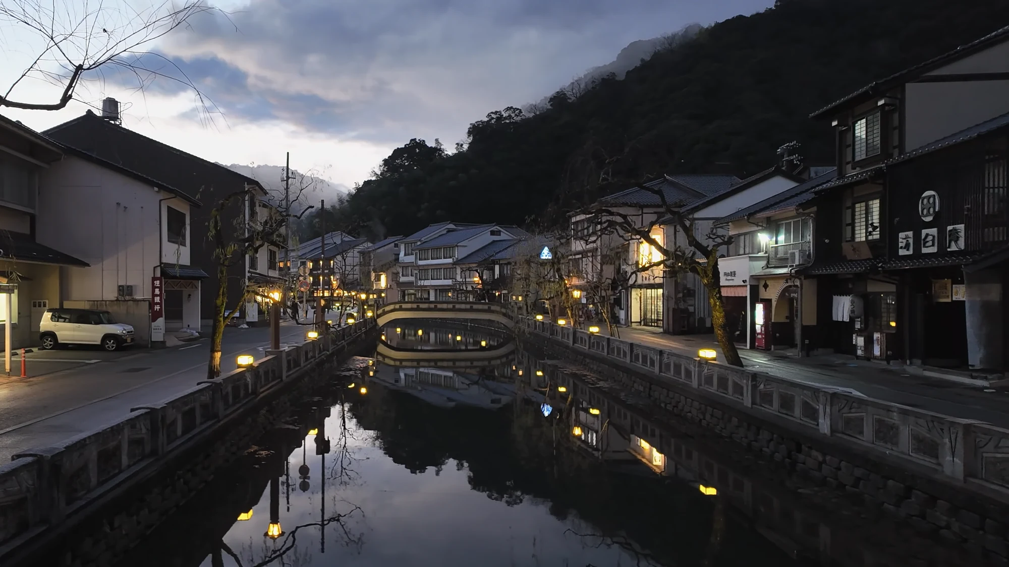 The Kinosaki canal at blue hour with lanterns and inn windows reflected in still water.