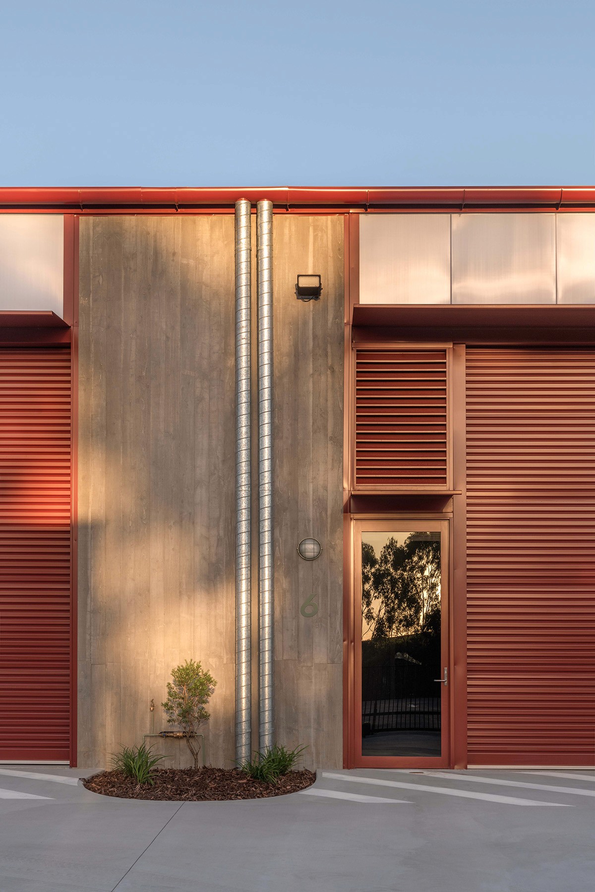 RedSheds façade detail showing a concrete wall with exposed vertical services, flanked by red louvred roller doors and a glazed pedestrian entry reflecting the surrounding landscape.
