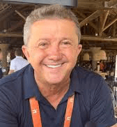 A person with curly hair and glasses smiles at the camera, seated in a cozy workspace with warm lighting.