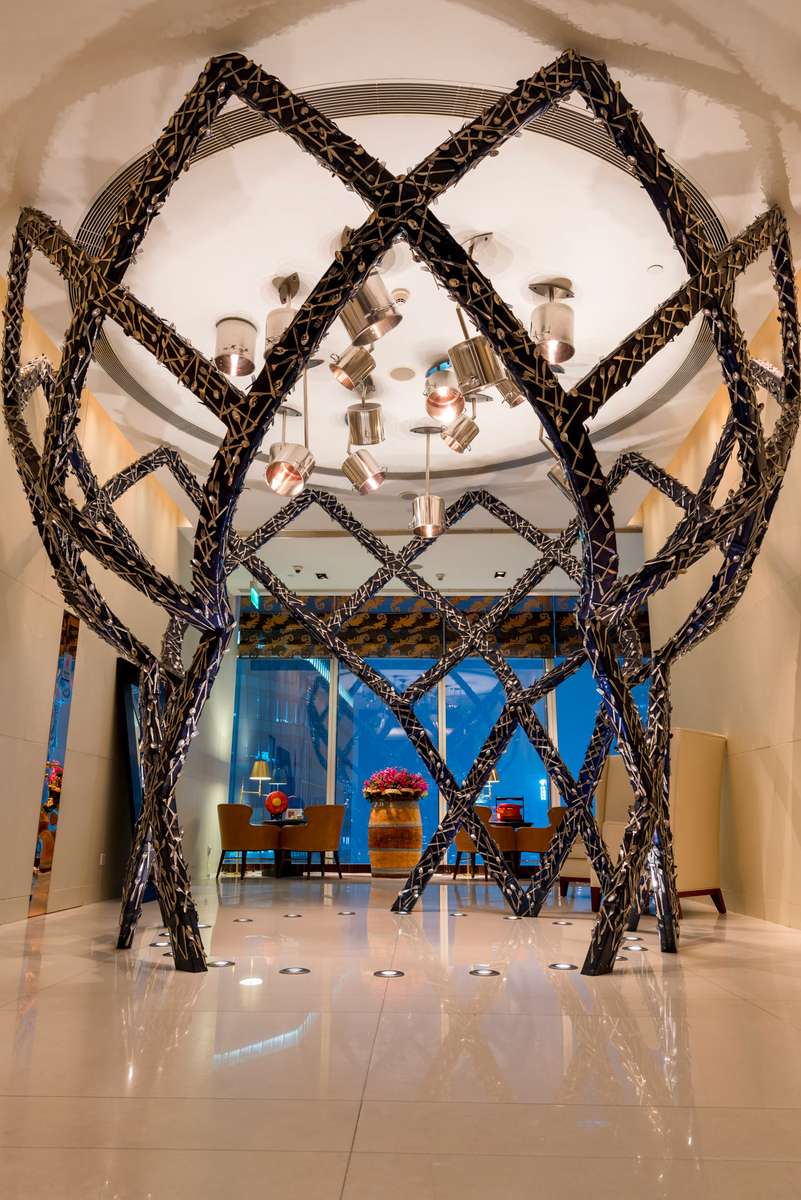A large, dark metal sculpture shaped like interlocking loops stands in a lobby with blue walls and polished tile floors.