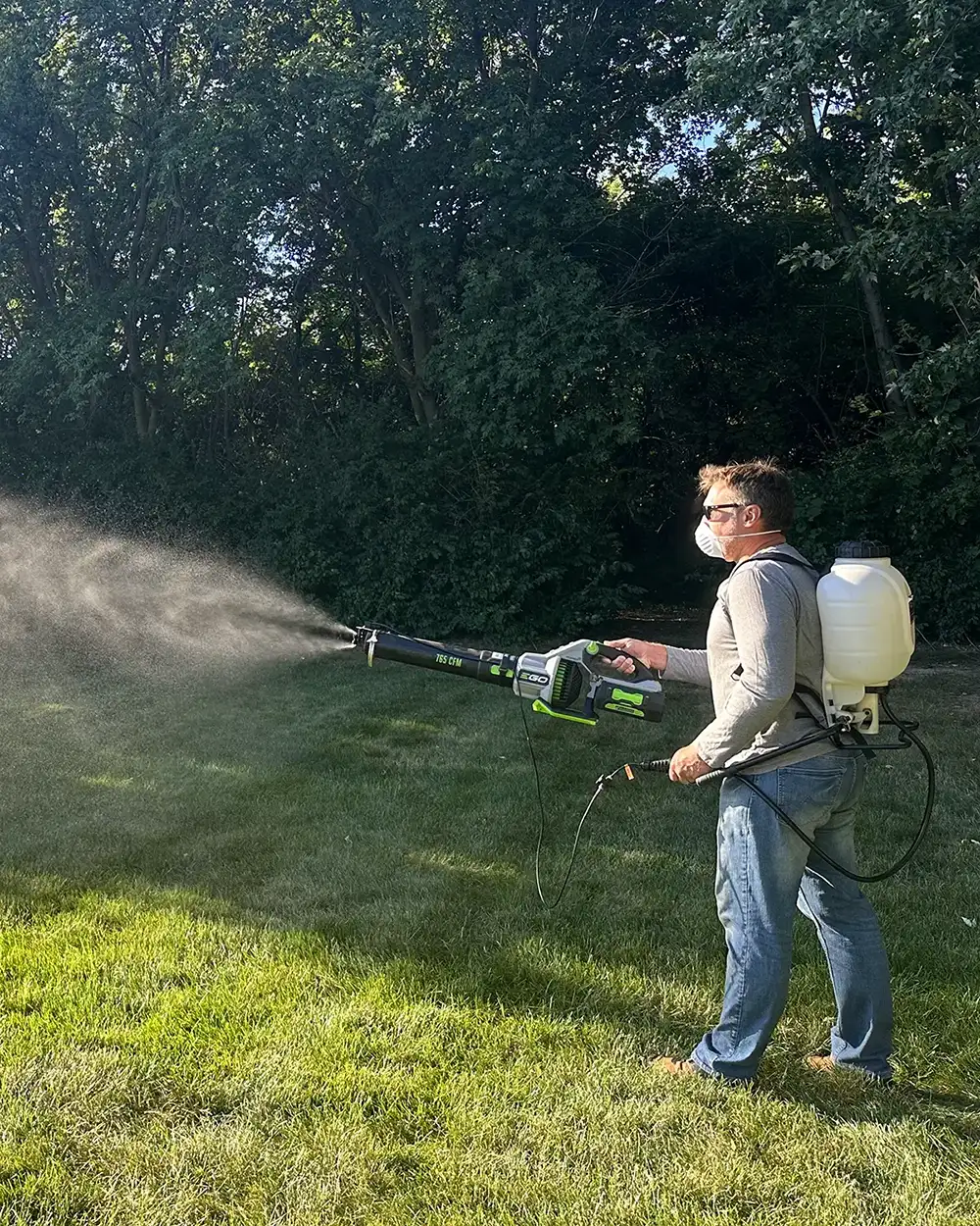 A person wearing a backpack sprayer applies a liquid spray in a green area under bright sunlight.