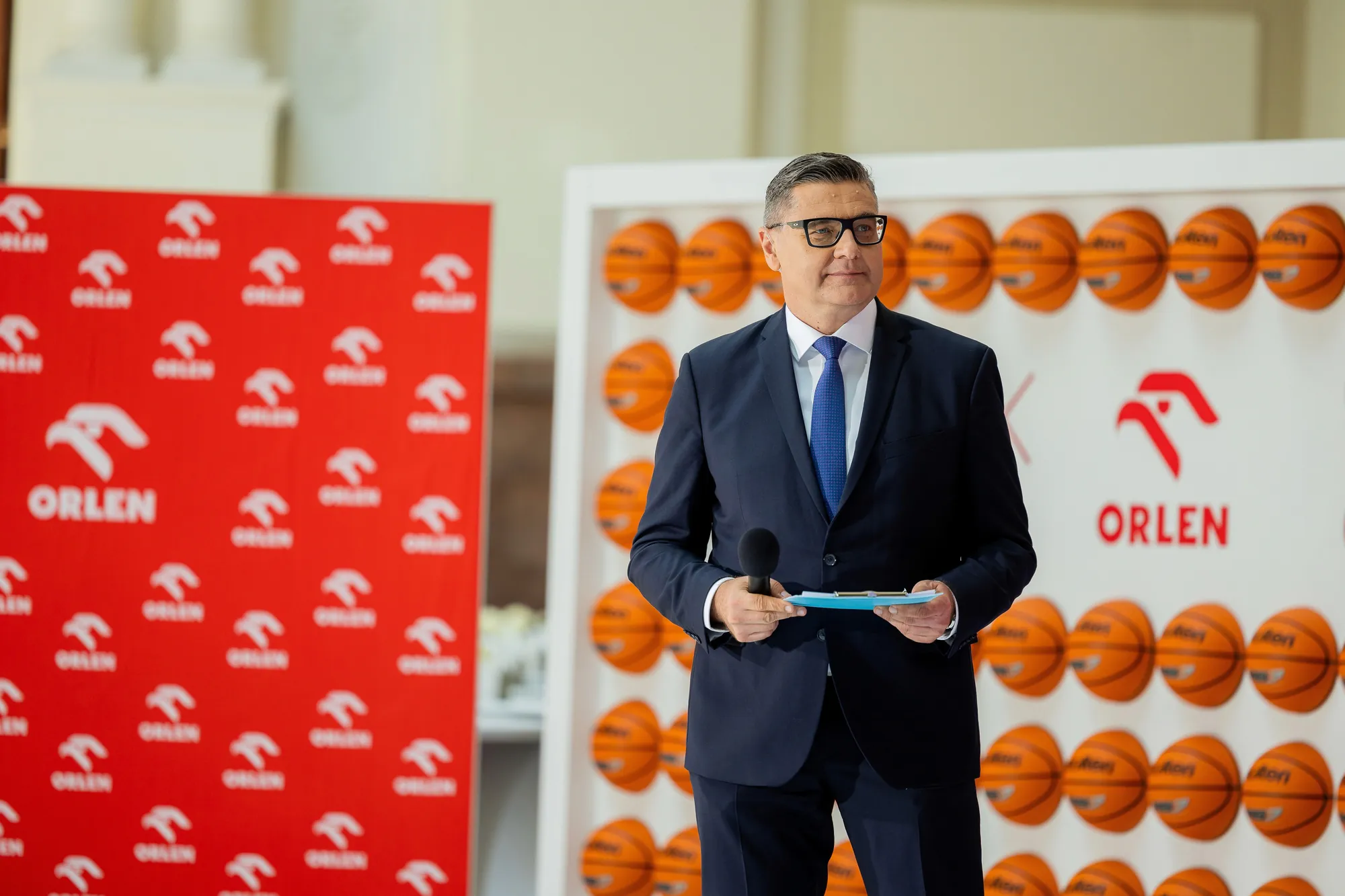 PZKosz i ORLEN – prowadzący konferencję prasową przed ścianką z piłkami koszykarskimi.