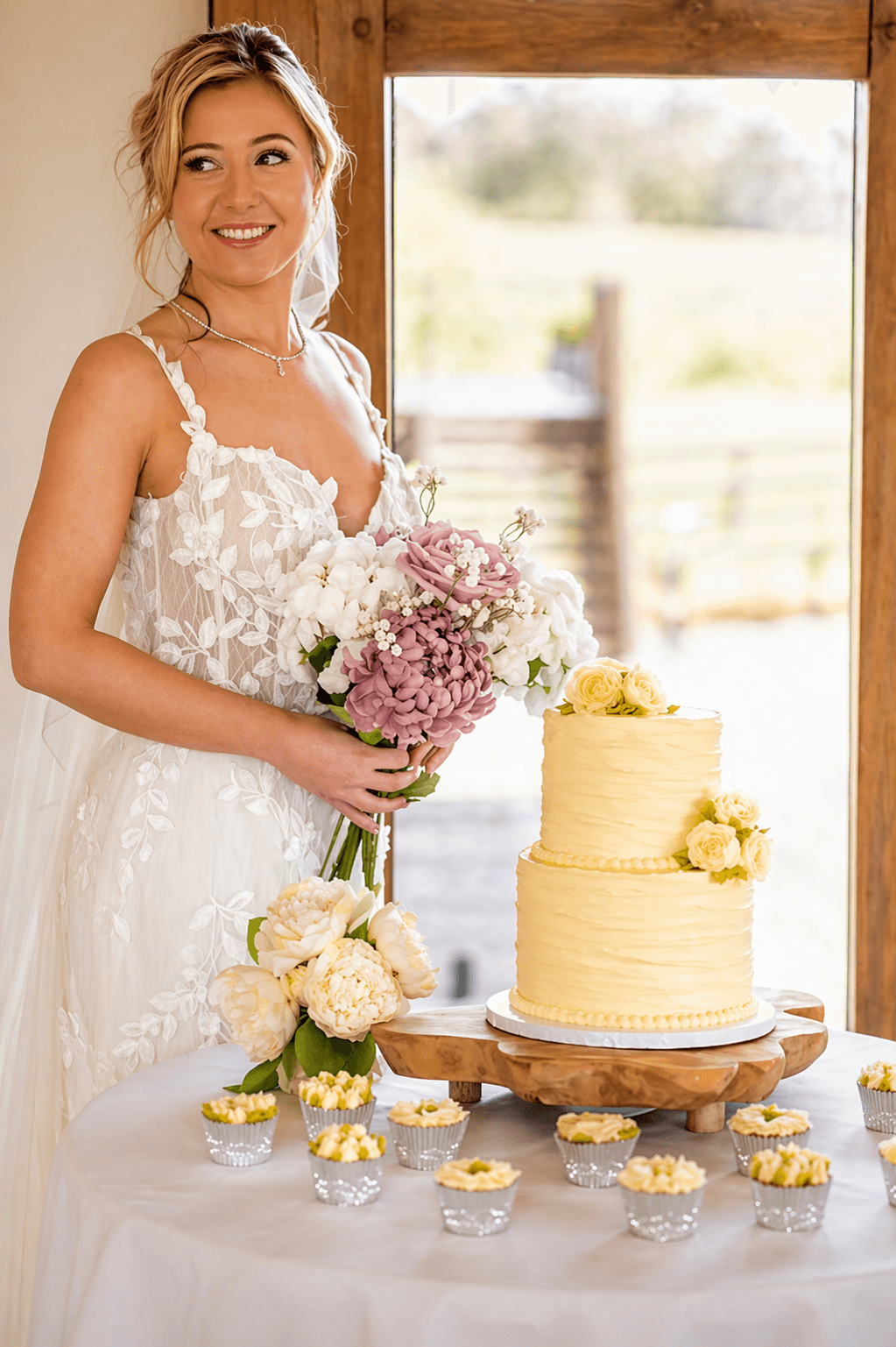 Bride holds bouquet & smiles beside a yellow cake and cupcakes on a white tablecloth, near a window.