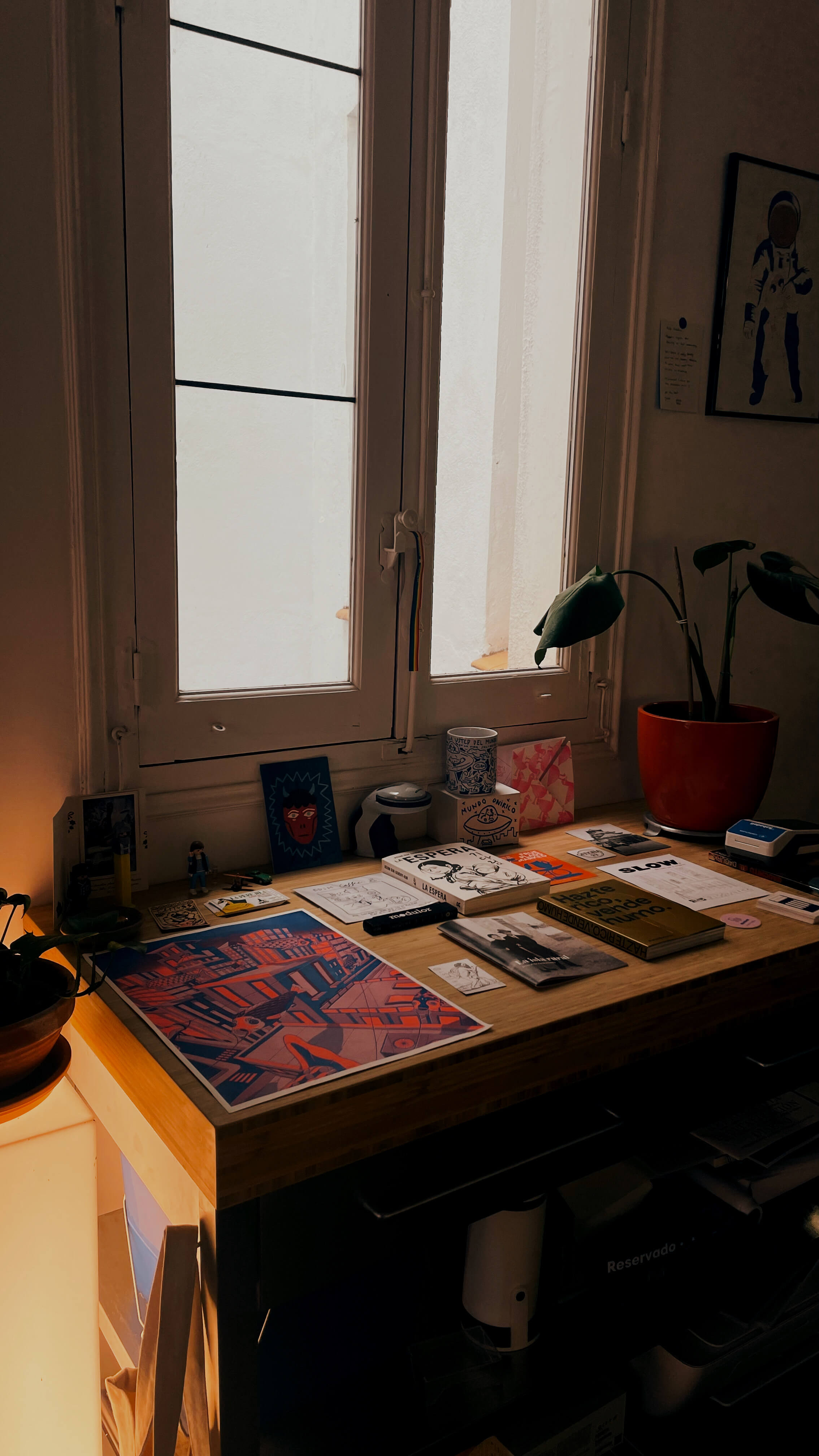 A dimly lit desk covered with art prints in front of a window