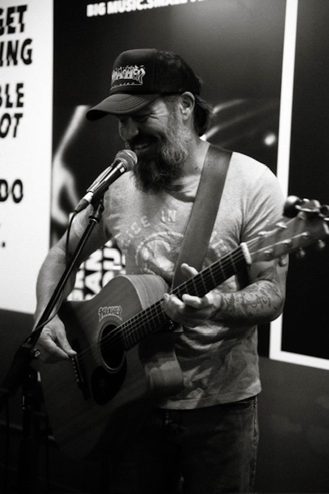 Scott Lennox smiles while performing solo with an acoustic guitar and microphone.