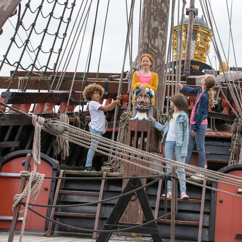 四个孩子站在一艘古老船只的甲板上，周围是绳索和木制结构。