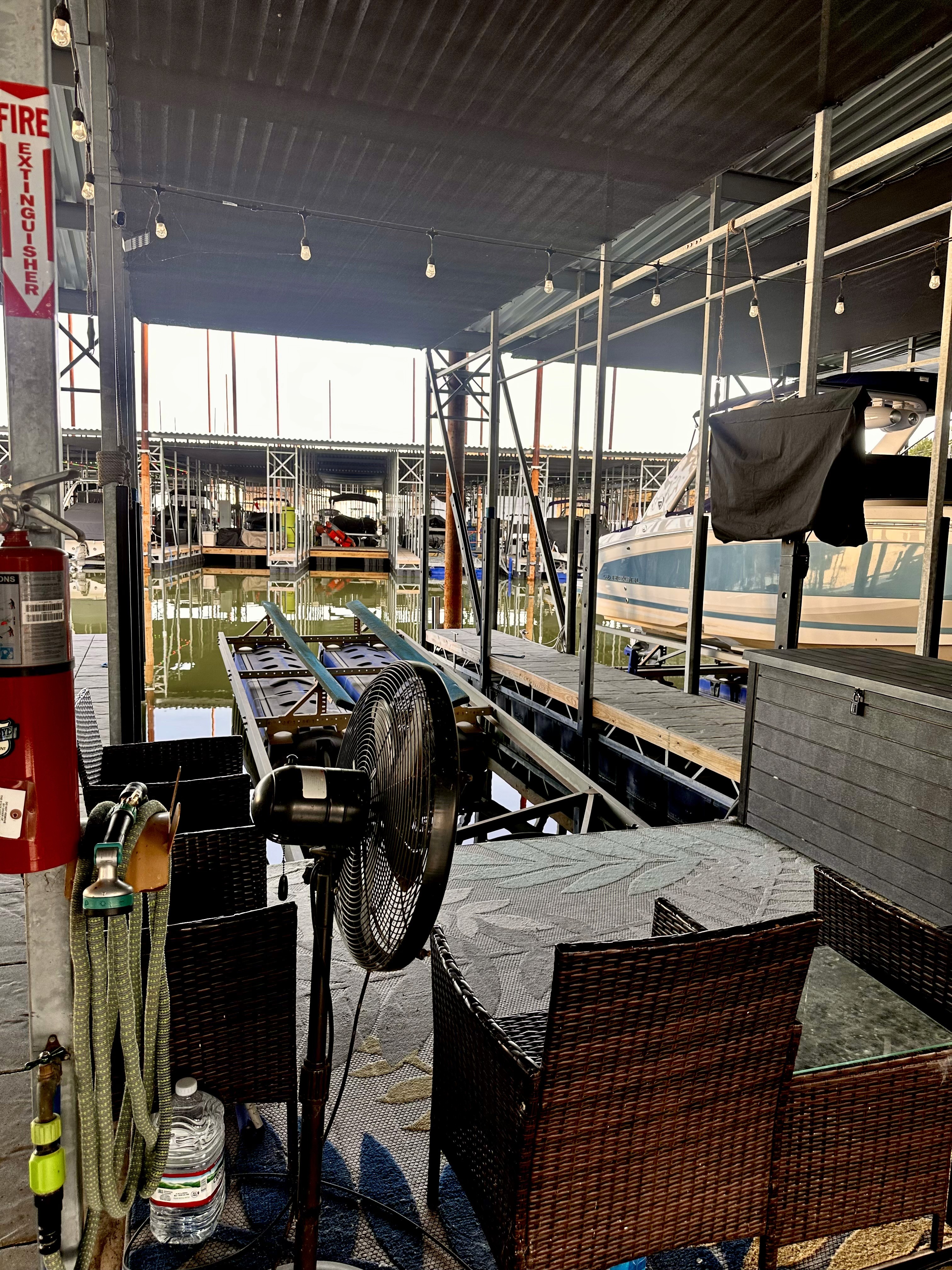 A cozy marina dock setup featuring a wicker patio dining set with a stone tabletop, a ceiling fan, and a fire extinguisher, overlooking boats moored in the calm waters under a covered structure with hanging lights.