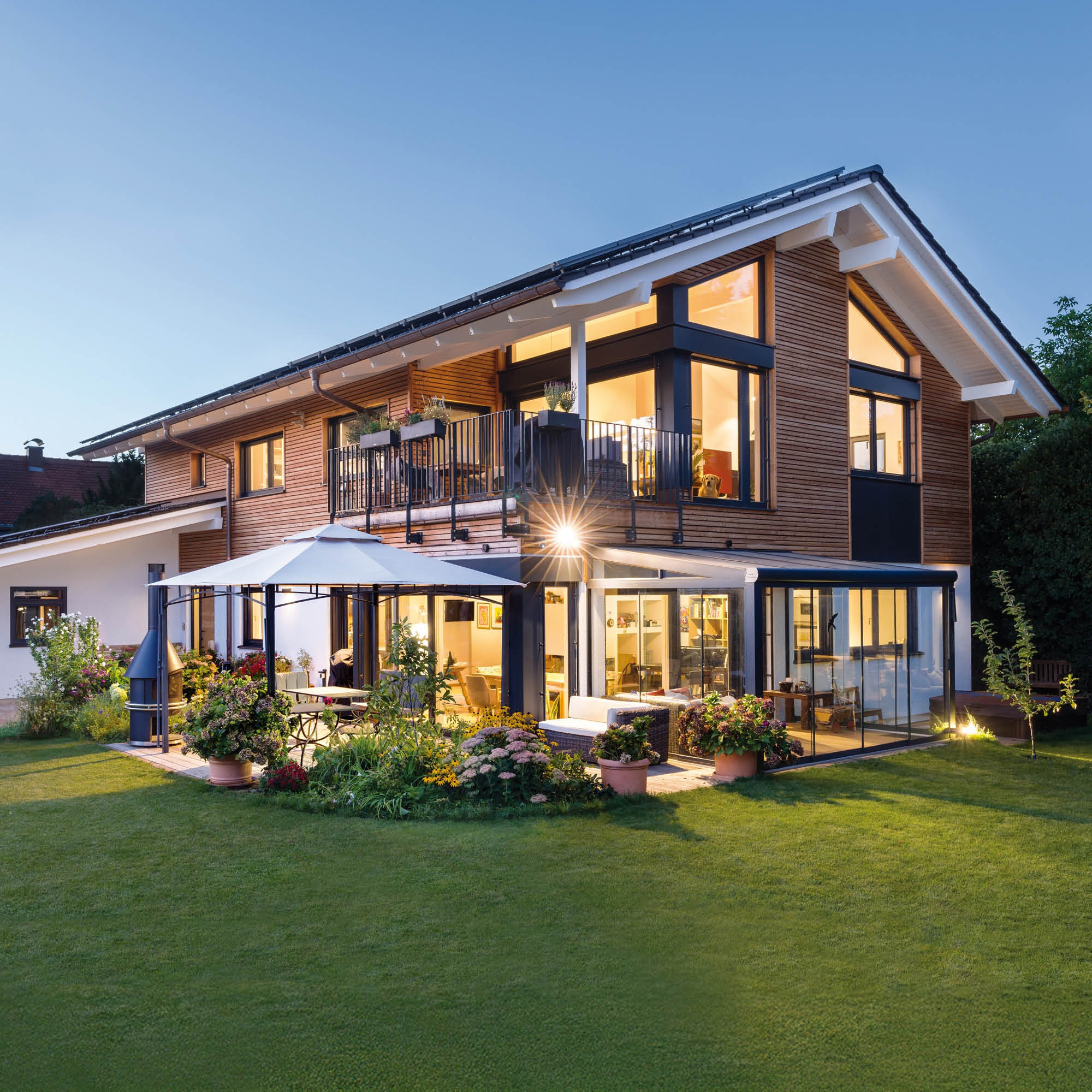 Modernes Holzhaus mit großzügiger Glasfassade, Balkon, Terrasse und Garten im Abendlicht.