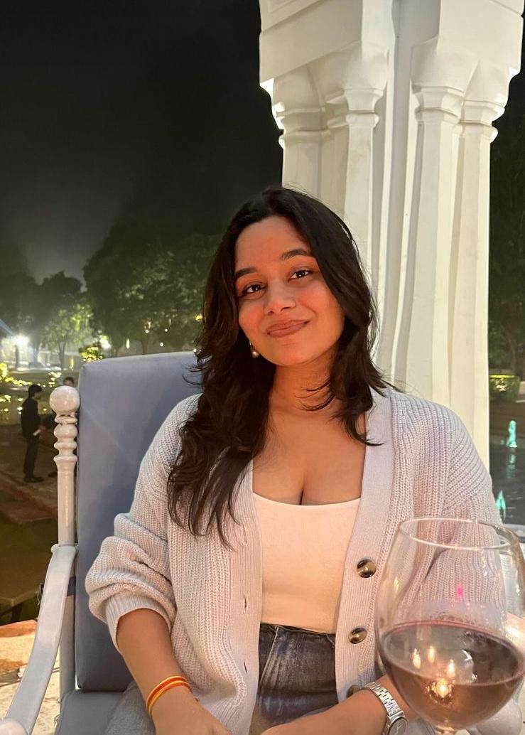 A person with short brown hair looks confidently at the camera, wearing a light pink blazer and white shirt. Soft lighting and plant in the background add a calming tone.