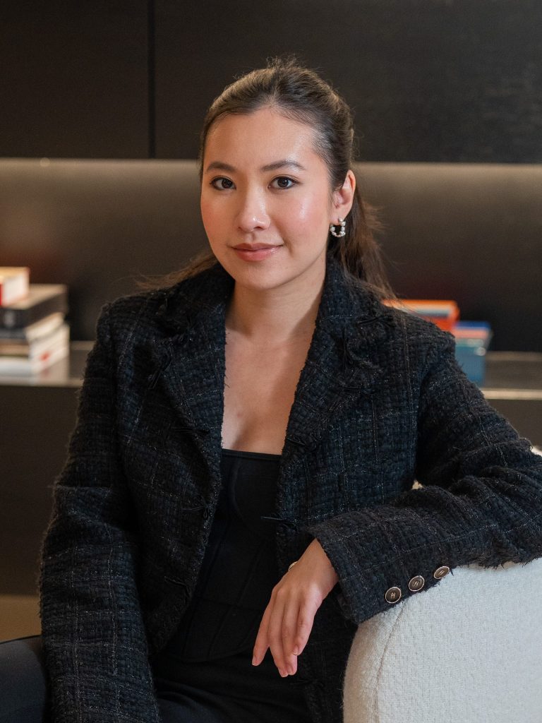 A portrait of a woman sitting on a couch, dressed in a black jacket, with her hand resting on her knee and a confident expression.